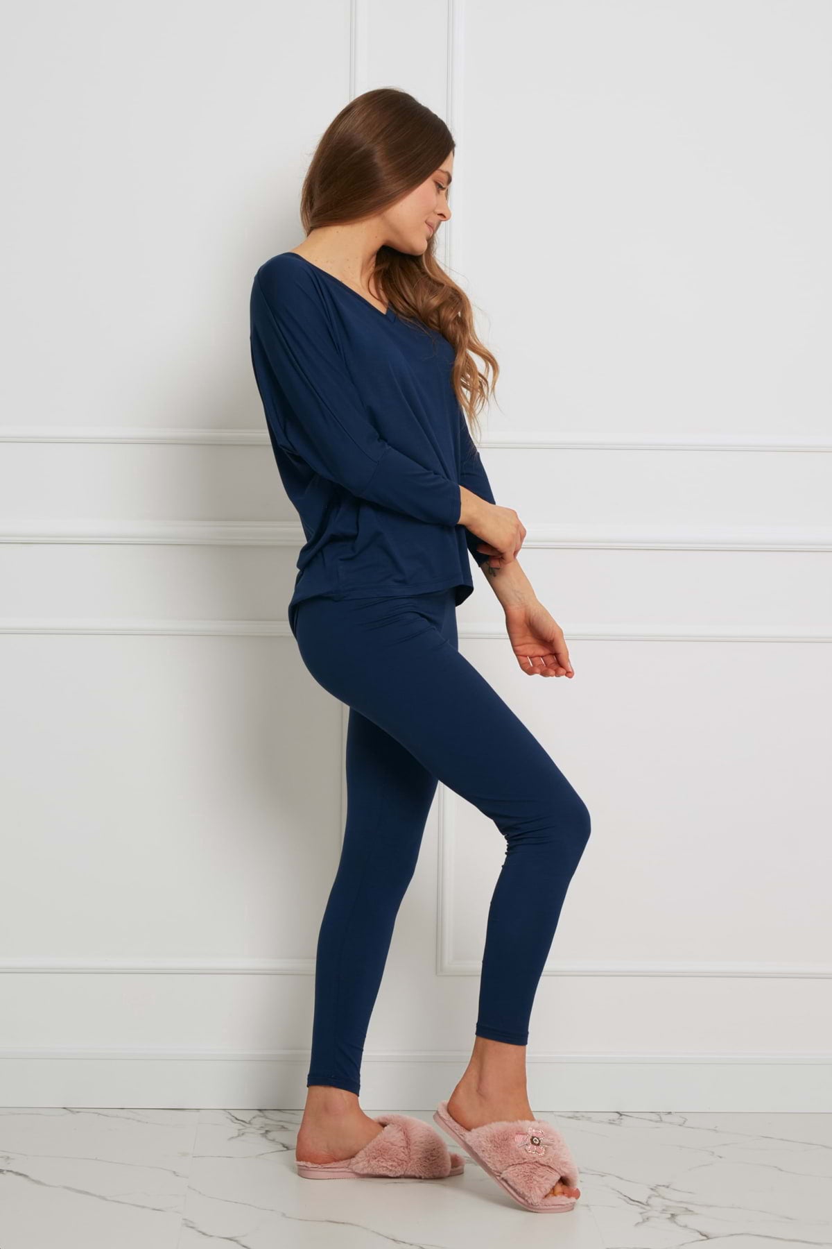 Kobieta z długimi brązowymi włosami stoi bokiem w pokoju, ubrana w Loungewear wiskozowy komplet z legginsami - granatowy z długimi rękawami i pasującymi legginsami, w połączeniu z jasnoróżowymi, rozmytymi kapciami. Spogląda w dół, jedna ręka zgięta w poprzek jej ciała.