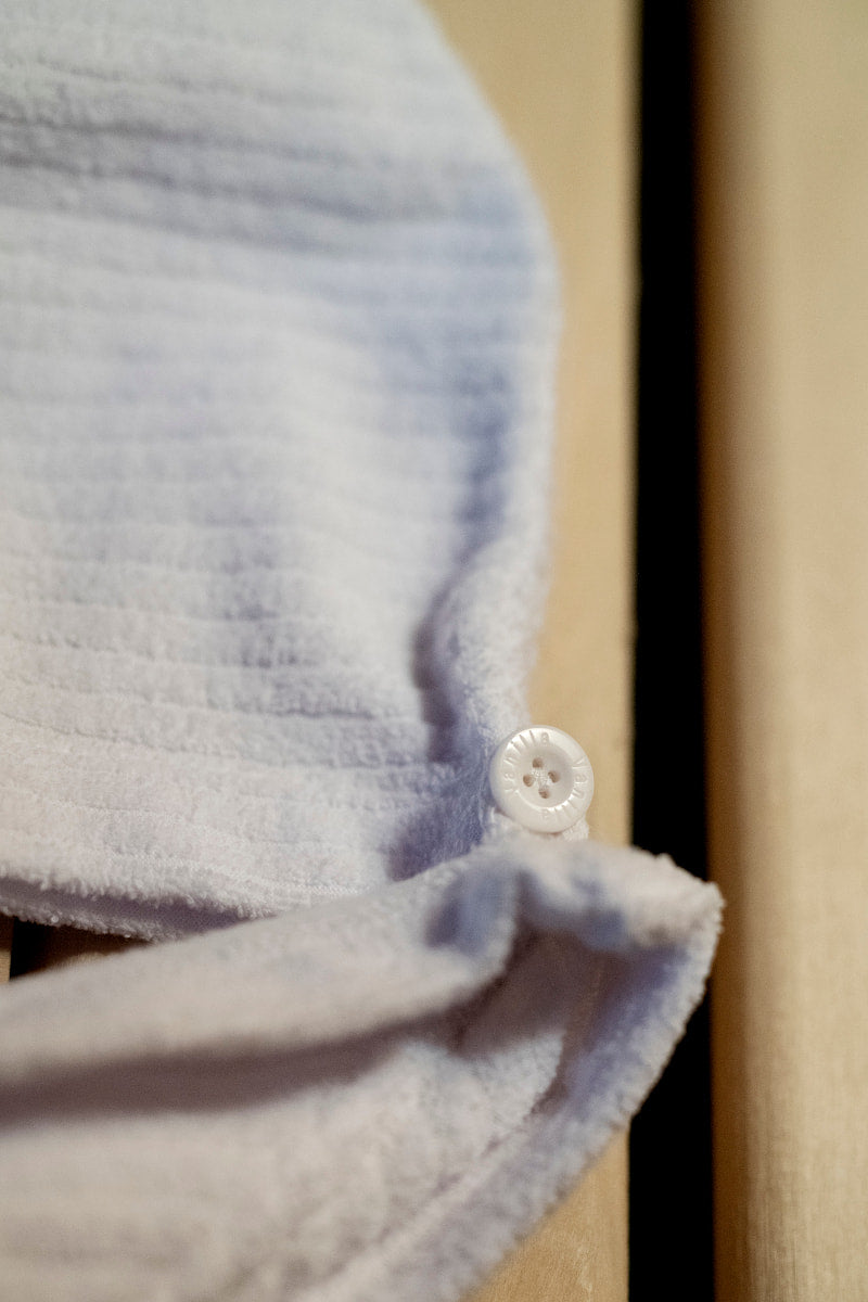 Zbliżenie na bawełniany turban Calma do włosów kąpielowy z guziczkiem - biały, wykonany z teksturowanej dzianiny frotte z małym zapięciem guziczkowym, pokazany na jasnej drewnianej powierzchni.