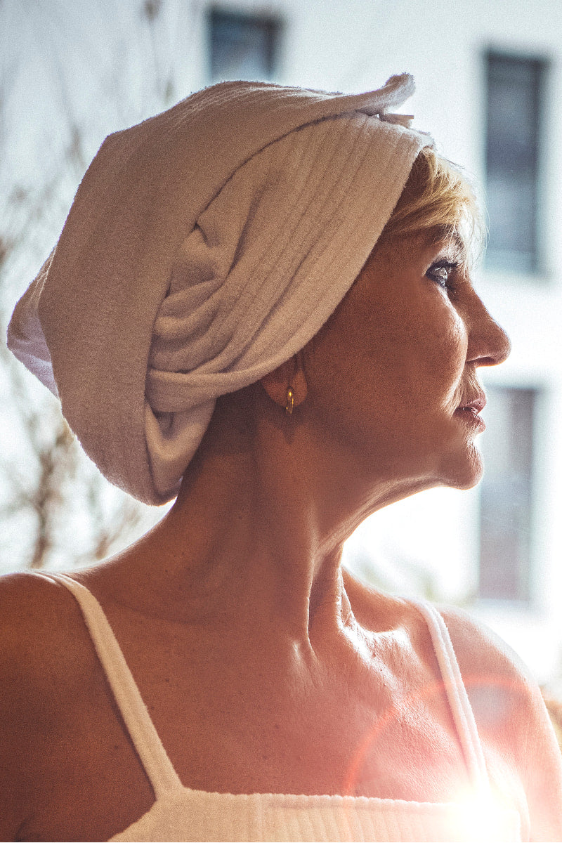 Kobieta z profilu nosi bawełniany turban Calma do włosów kąpielowy z guziczkiem - biały i top bez rękawów, stojąc przy oknie z promieniami słońca na ramieniu.