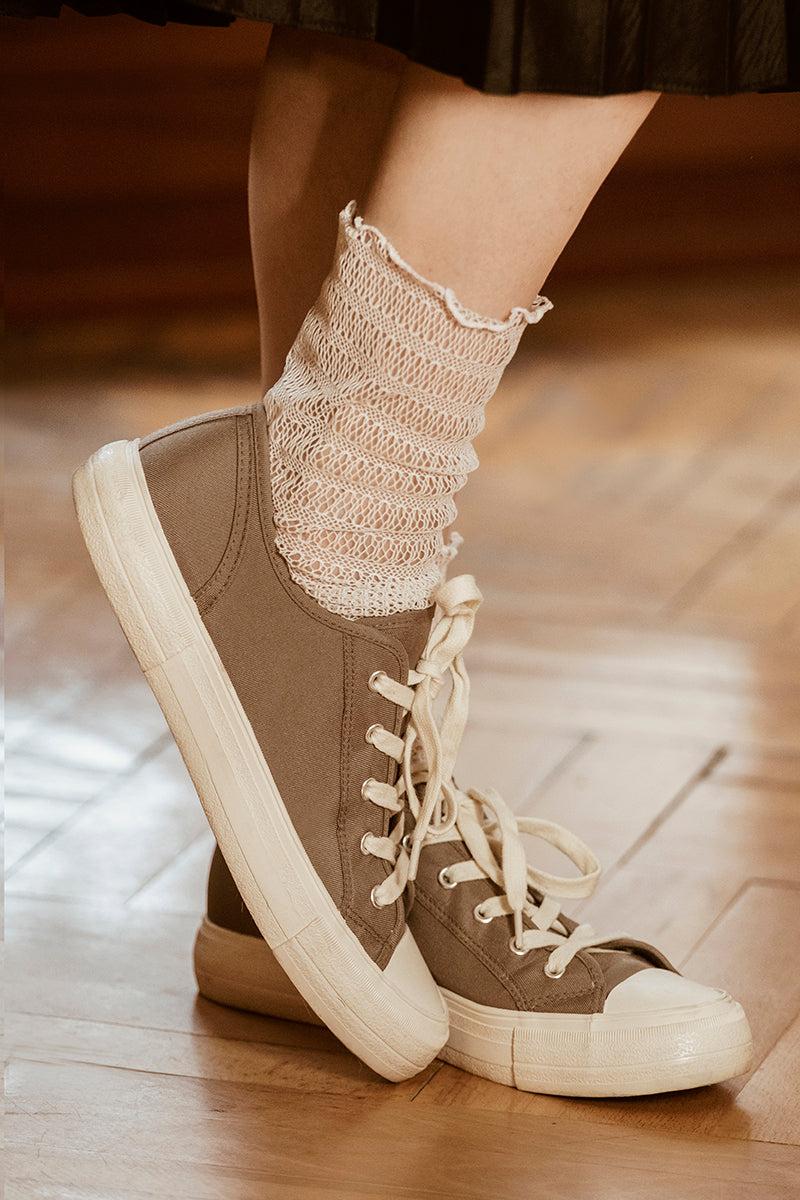 Osoba w kabaretkach Skarpetki - Waniliowa Sieć i płóciennych trampkach w kolorze taupe z białymi podeszwami stoi na drewnianej podłodze, jedna stopa lekko uniesiona i skrzyżowana - subtelny ukłon w stronę eleganckich stylizacji.