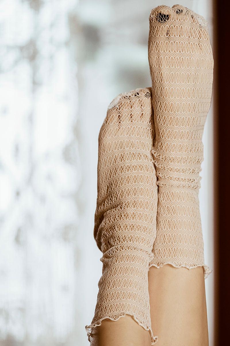 Dwie stopy w kabaretkach Skarpetki - Waniliowa Sieć, z widocznymi dziurami na palcach, spoczywają na rozmytym tle, wprowadzając wyjątkowy akcent do eleganckich stylizacji.