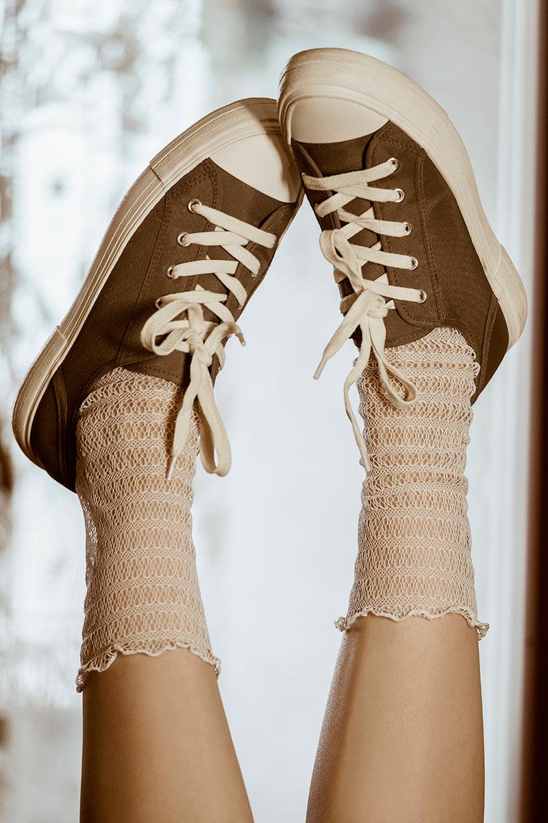 Dwie nogi uniesione w powietrzu noszą brązowe sznurowane trampki i kabaretki Skarpetki - Waniliowa Sieć. Delikatnie rozmyte tło dodaje przytulnego, zabawnego klimatu, nawiązując do eleganckich stylizacji.