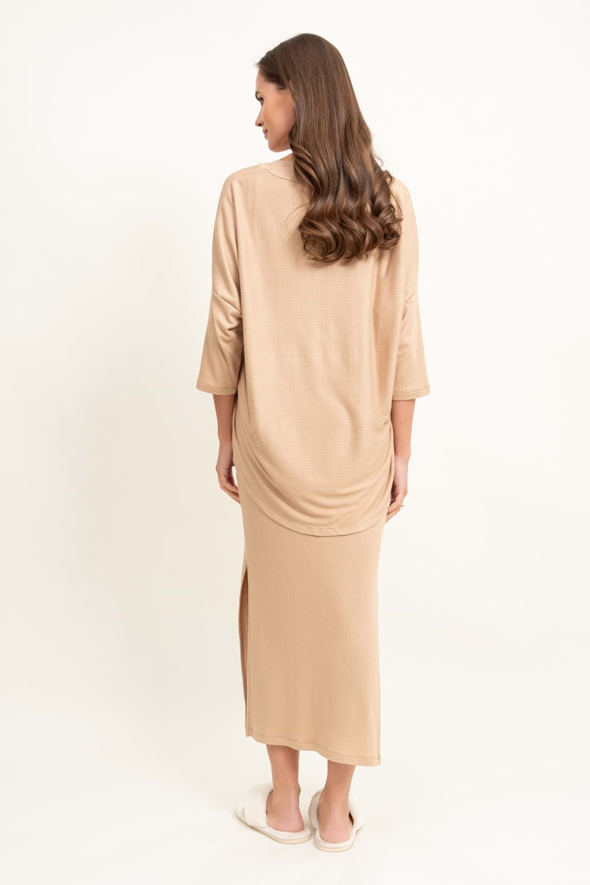 Kobieta z długimi brązowymi włosami stoi odwrócona, ubrana w Loungewear bluzka oversize z asymetrycznymi cięciami - wielbłąd i białe pantofle na prostym, jasnym tle.