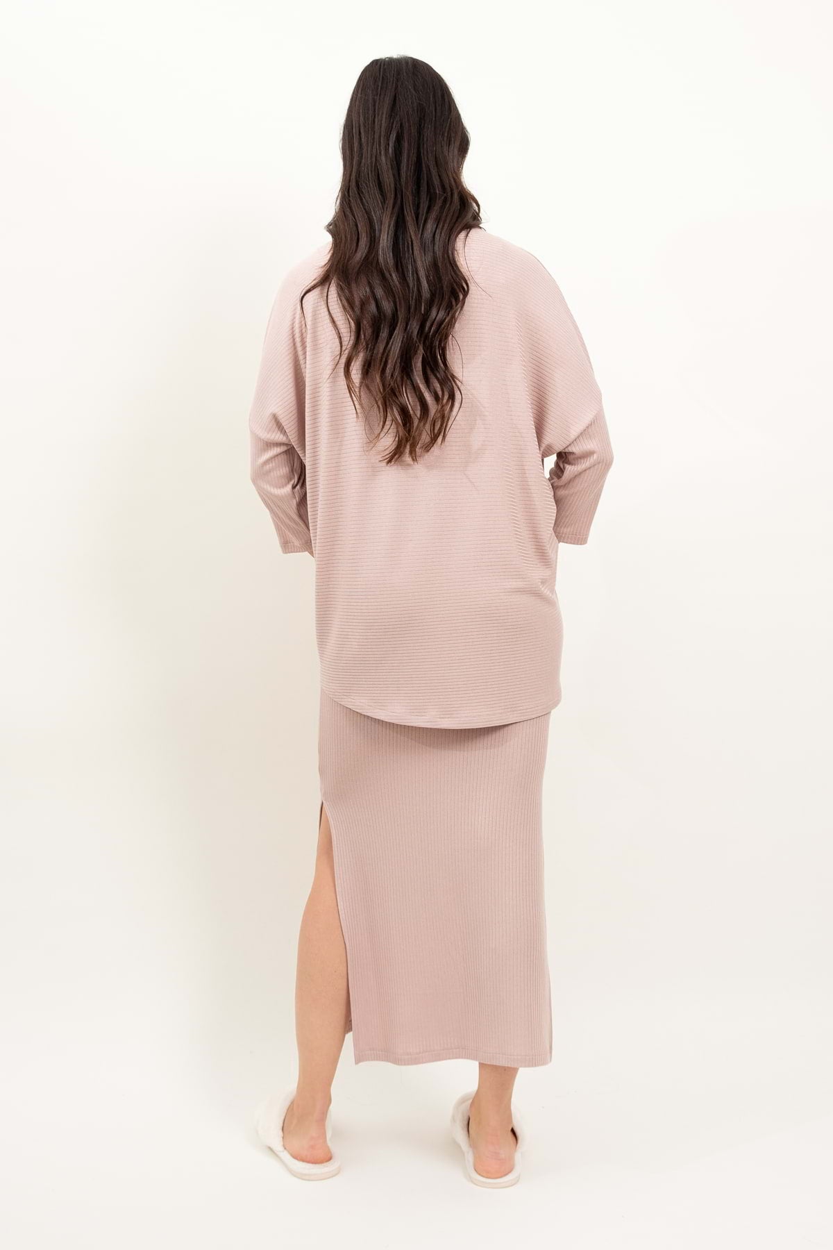 Kobieta z długimi falistymi brązowymi włosami stoi odwrócona, ubrana w pudrowy róż loungewear bluzka oversize z asymetrycznymi cięciami. Jest boso na prostym, jasnym tle.