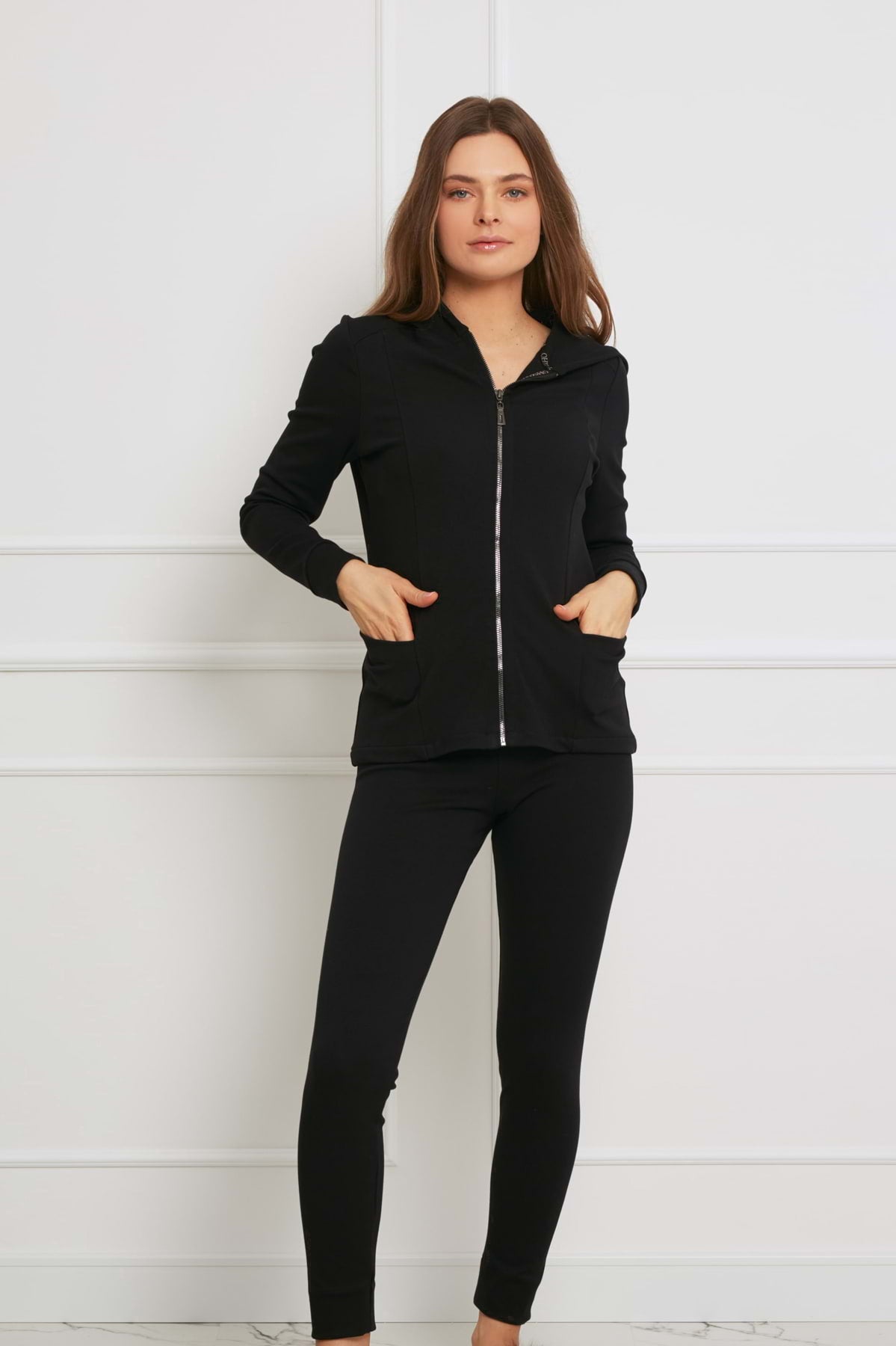 Kobieta stoi w pomieszczeniu na tle białej ściany, ubrana w elegancki strój: Loungewear dopasowana bluza bawełniana z kapturem - czarna i dopasowane czarne legginsy, ręce w kieszeniach kurtki i neutralny wyraz twarzy.