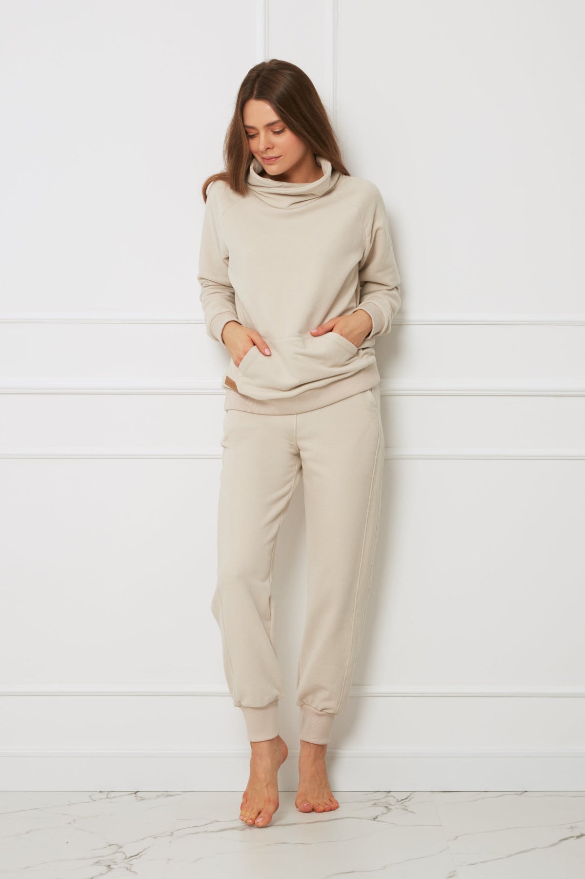 Kobieta, boso na tle białej ściany, ma na sobie dopasowaną jasnobeżową bluzę z dekoltem i Loungewear miękkie spodnie bawełniane - beżowe. Z rękami w kieszeniach spogląda w dół ze zrelaksowanym wyrazem twarzy.