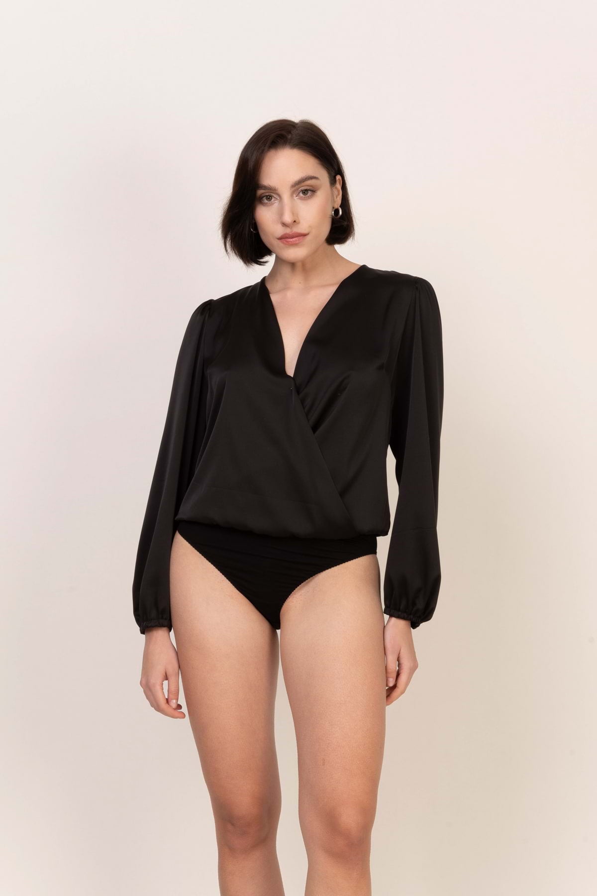 Kobieta o krótkich ciemnych włosach ma na sobie Loungewear satynowa bluzka-body z długim rękawem - czarna z eleganckimi bufiastymi rękawami i dekoltem w szpic, w połączeniu z czarnymi spodniami z wysokim stanem na jasnym tle.