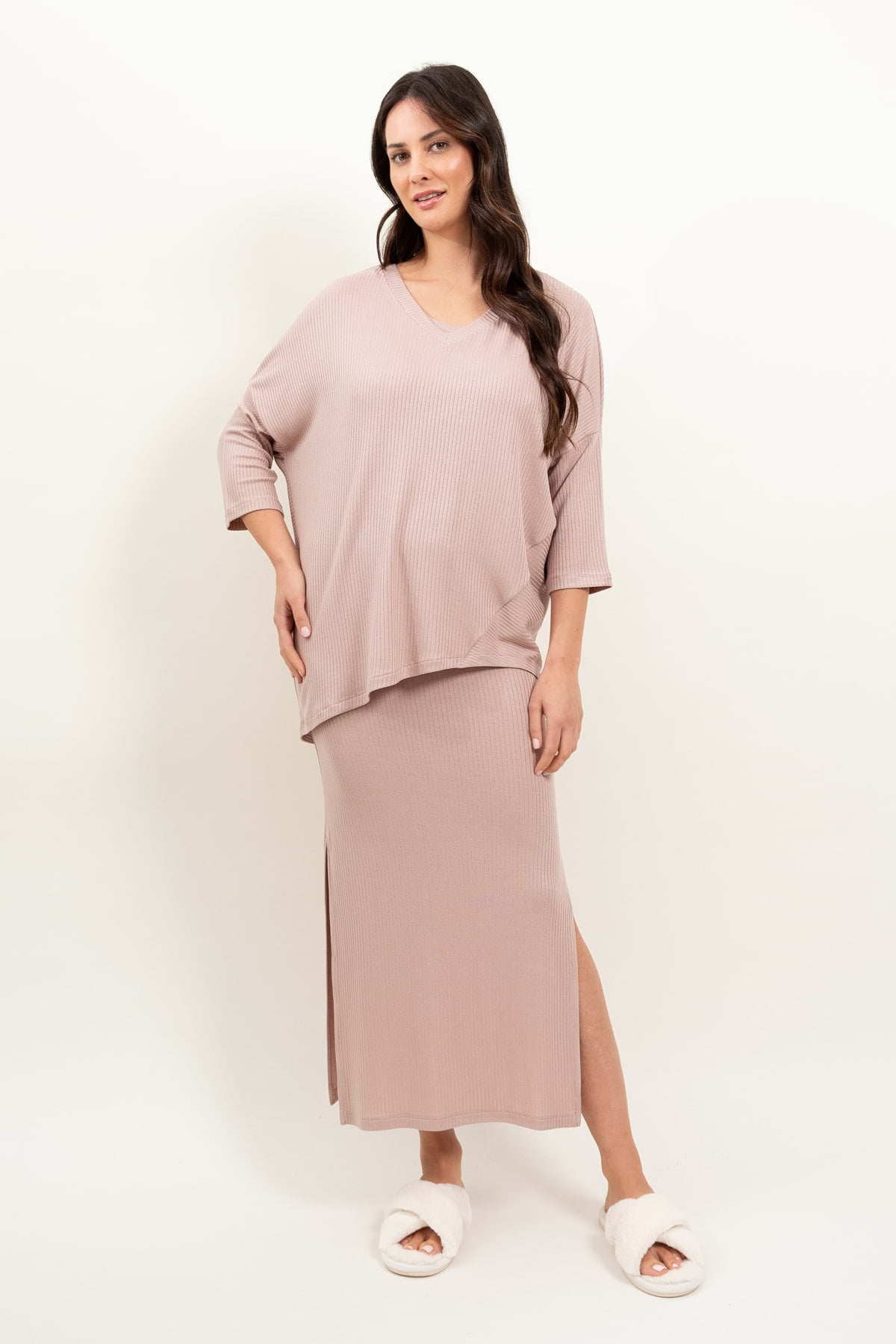 Kobieta z długimi, falowanymi brązowymi włosami pozuje w zrelaksowanym nastroju na białym tle, ubrana w bluzkę oversize Loungewear z asymetrycznymi cięciami - pudrowy róż i koordynującą prążkowaną spódnicę midi z rozcięciem z boku oraz białe, rozmyte kapcie.