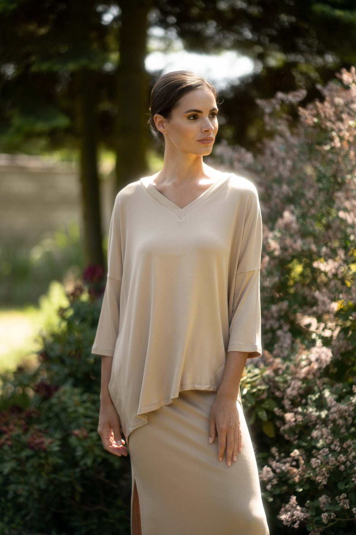 Kobieta stoi na zewnątrz w delikatnym słońcu, ubrana w Loungewear bluzka oversize z asymetrycznymi cięciami - camel i pasującą spódnicę, otoczona bujną zielenią i kwitnącymi krzewami, patrząc spokojnie w bok.