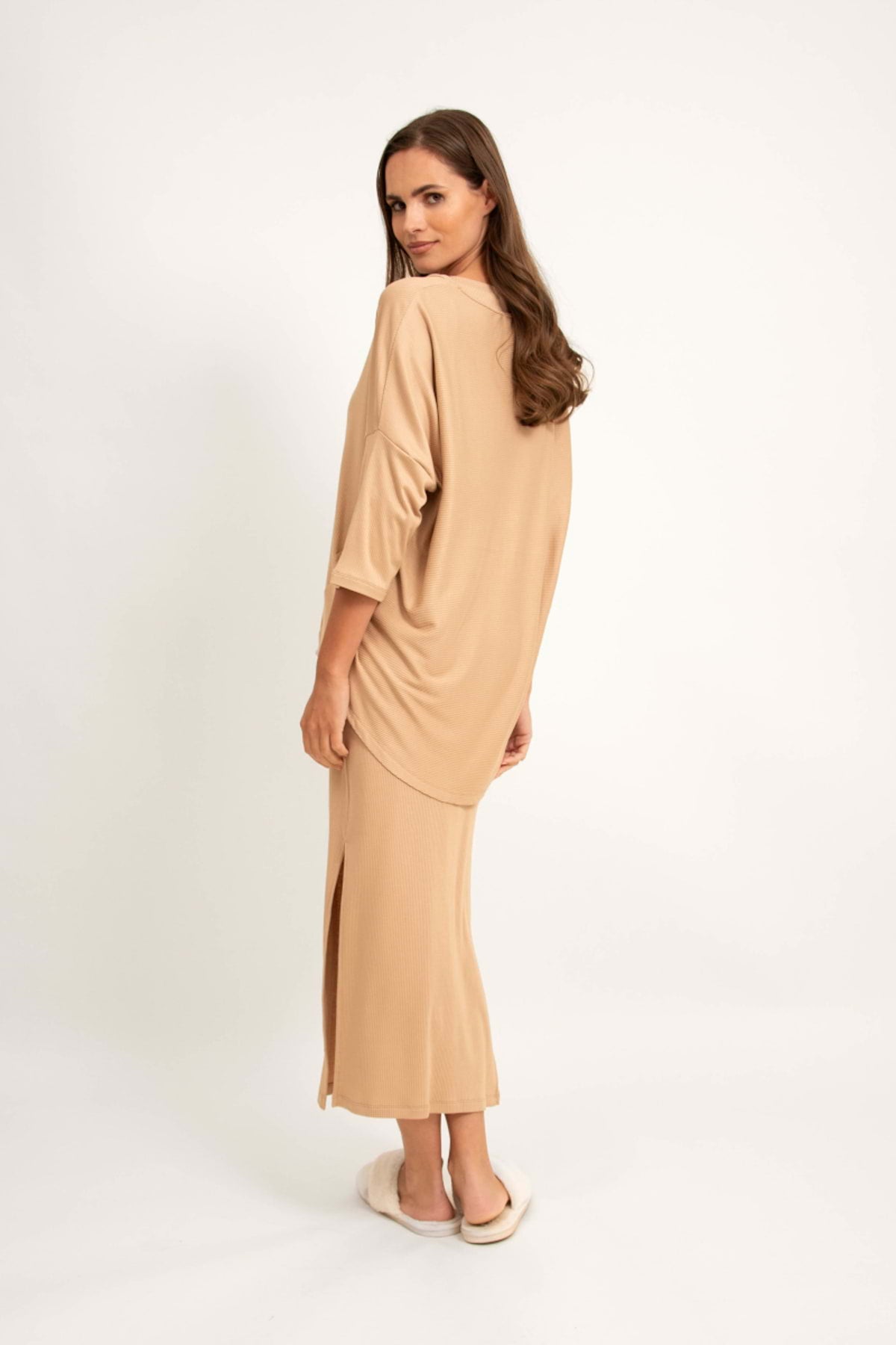 Kobieta z długimi brązowymi włosami ma na sobie Loungewear bluzkę oversize z asymetrycznymi cięciami - camel i dopasowaną spódnicę do kostek, stojąc boso, odwrócona na bok i patrząc przez ramię na jednolitym jasnym tle.