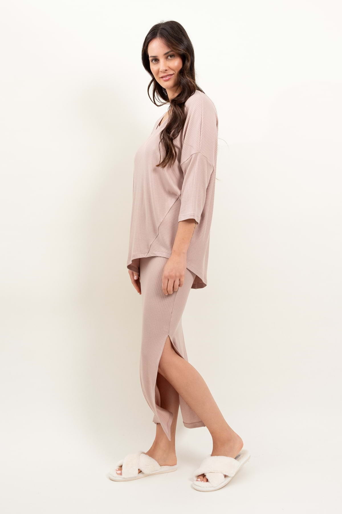 Kobieta z długimi brązowymi włosami nosi Loungewear bluzka oversize z asymetrycznymi cięciami - pudrowy róż i dopasowaną spódnicę, stojąc bokiem i uśmiechając się w białych rozmytych kapciach na białym tle.