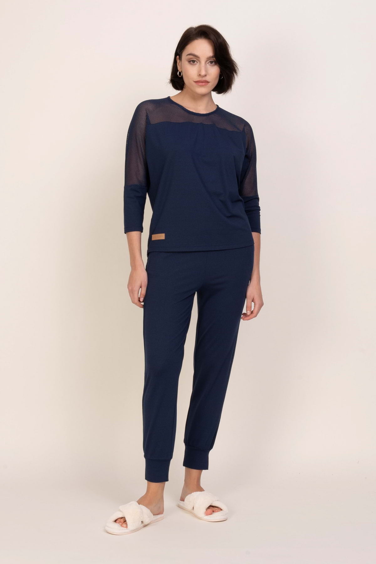 Kobieta z krótkimi ciemnymi włosami i neutralnym wyrazem twarzy stoi na prostym tle ubrana w zestaw "Loungewear wiskozowa bluzka z siateczką - ciemny jeans" z pasującymi spodniami i białymi rozmytymi kapciami.
