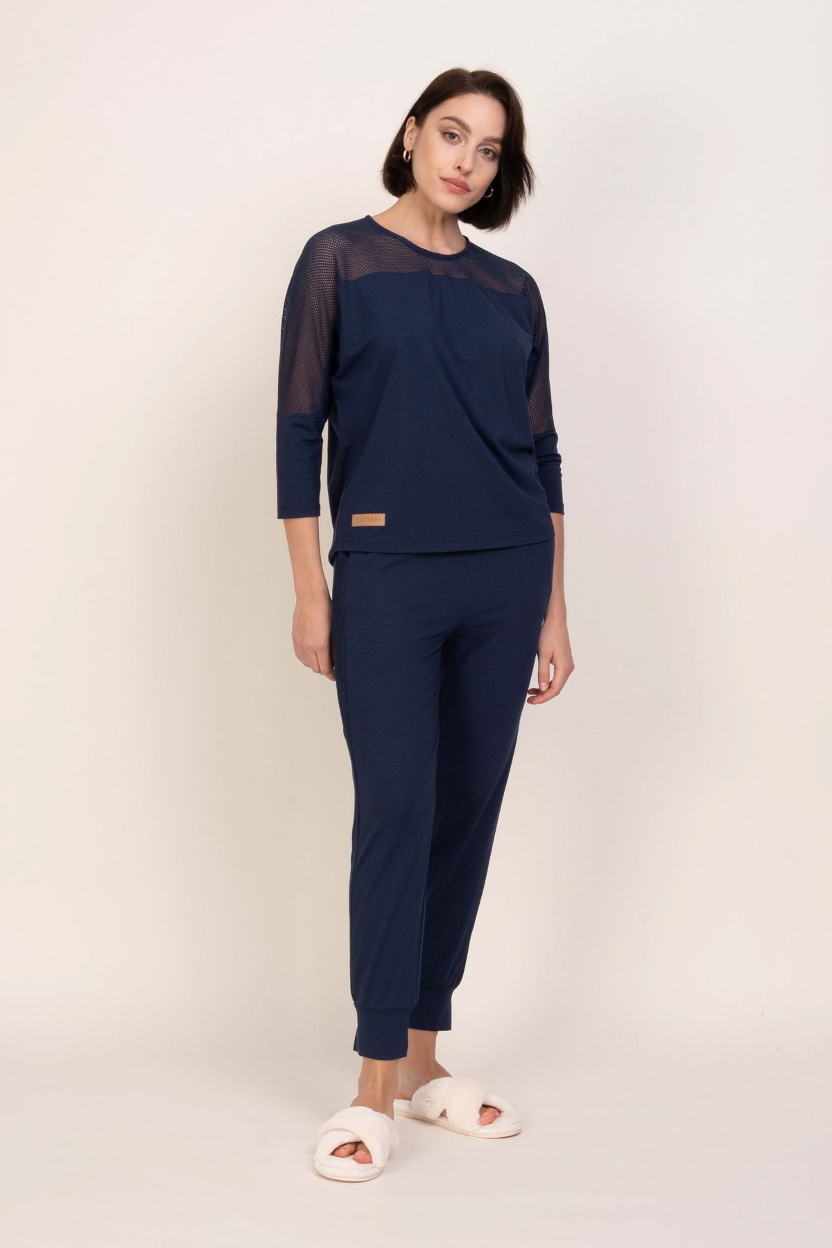 Kobieta z krótkimi brązowymi włosami i zrelaksowanym wyrazem twarzy stoi na prostym tle, ubrana w "Loungewear wiskozowa bluzka z siateczką - ciemny jeans" z dopasowanymi spodniami i białymi, rozmytymi kapciami.