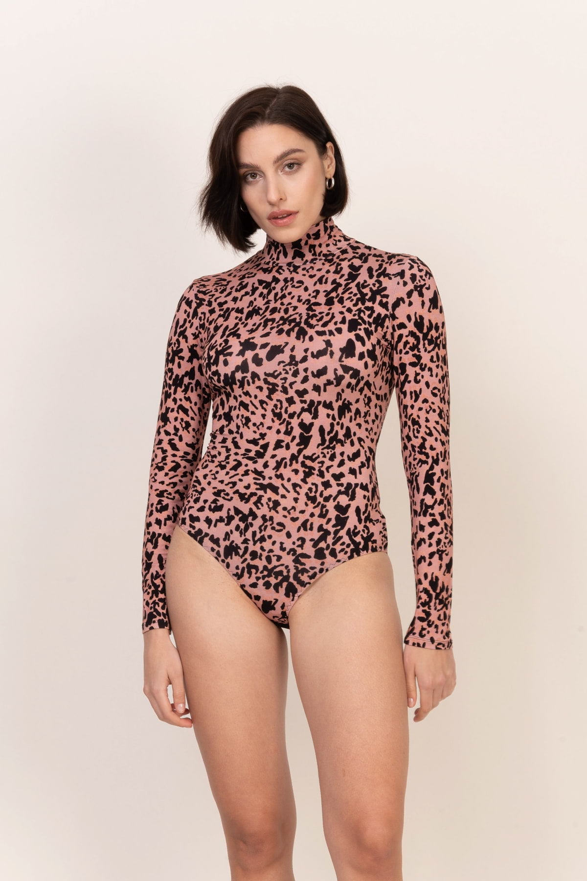 Kobieta z krótkimi brązowymi włosami nosi Loungewear wiskozowe body z golfem - panterka, z golfem i nadrukiem w panterkę. Stoi na jasnym tle, patrząc w kamerę z neutralnym wyrazem twarzy.