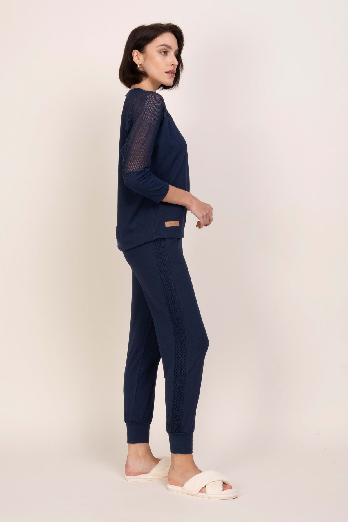 Kobieta o krótkich ciemnych włosach stoi z profilu na prostym tle, ubrana w zestaw "Loungewear wiskozowa bluzka z siateczką - ciemny jeans" i kremowe rozmyte kapcie, z rękami spoczywającymi w kieszeniach.