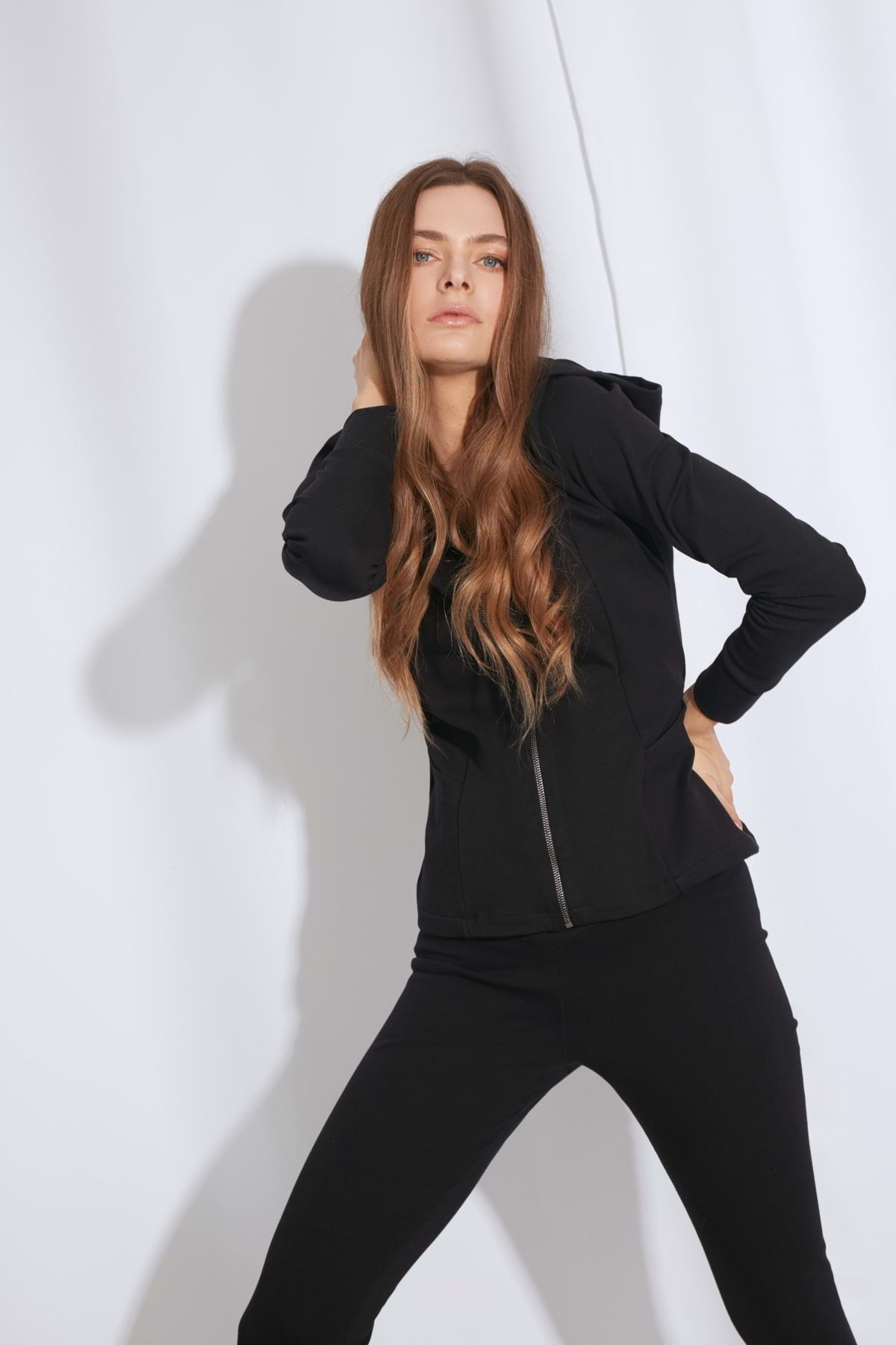 Kobieta o długich, falowanych brązowych włosach modeluje Loungewear dopasowana bluza bawełniana z kapturem - czarna i pasujące czarne spodnie, emanujące eleganckim stylem z grubej dzianiny na białym tle.