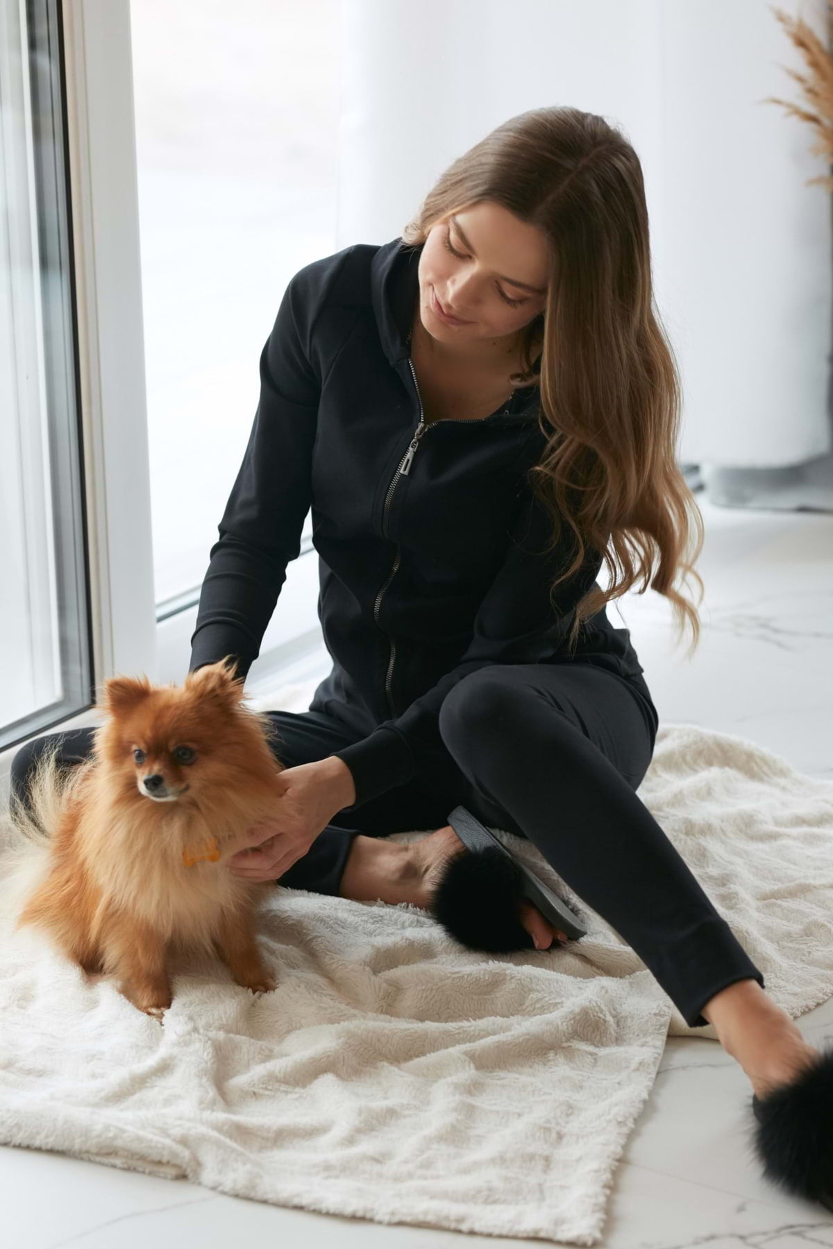 Kobieta w Loungewear dopasowane spodnie bawełniane - czarne siedzi na kocu przy oświetlonym słońcem oknie, delikatnie trzymając małego puszystego brązowego psa. Przytulny, jasny pokój ma białe podłogi i mnóstwo naturalnego światła.
