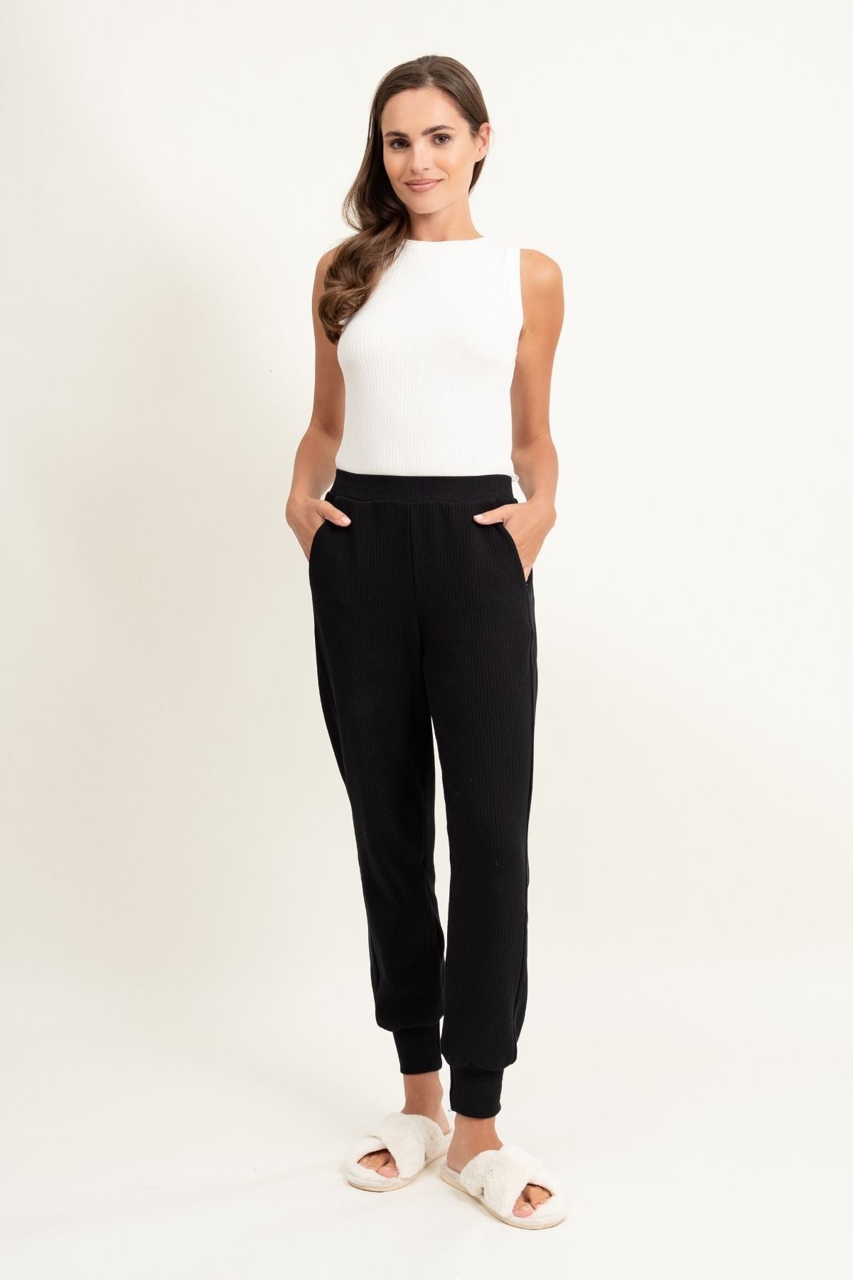 Kobieta stoi na białym tle, ubrana w spodnie dresowe Loungewear z lampasami - czarne, biały top bez rękawów i białe pantofle. Uśmiecha się z rękami w kieszeniach i ma długie brązowe włosy.