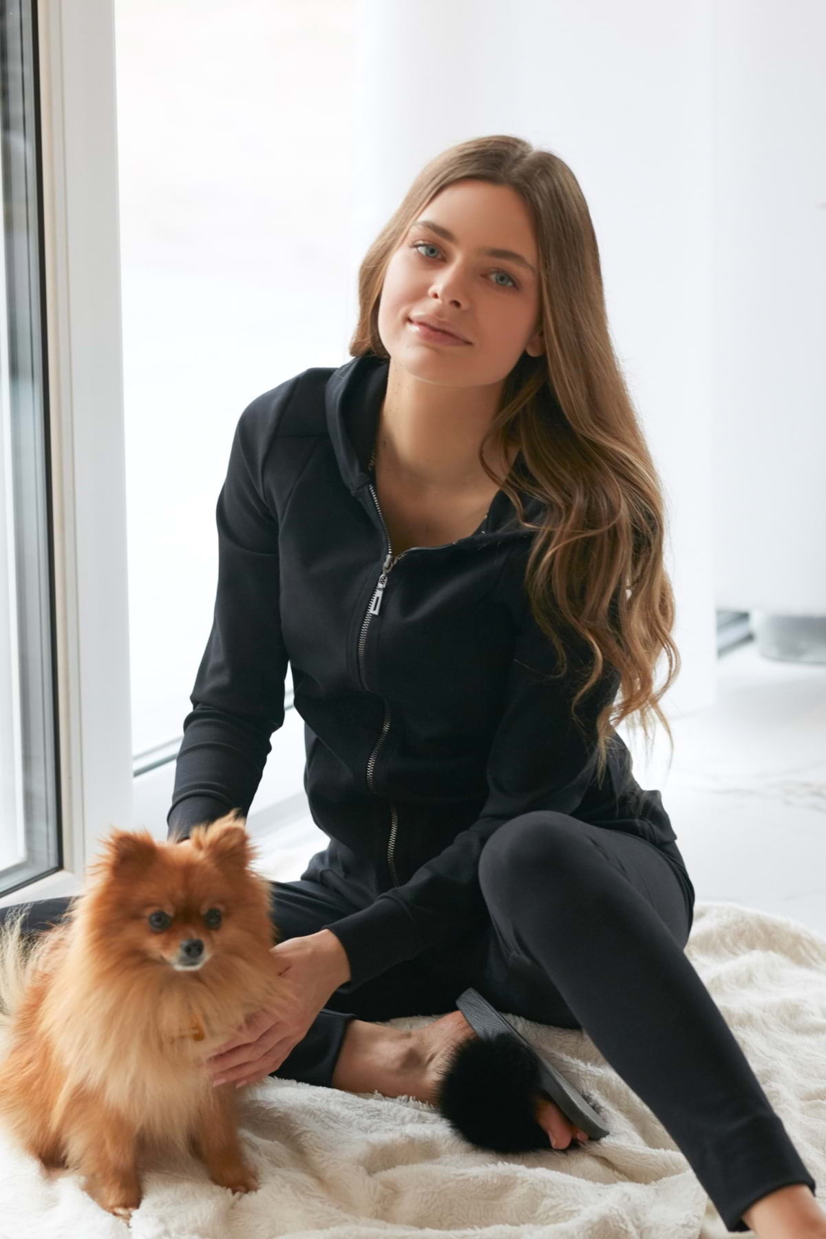 Kobieta w Loungewear dopasowana bluza bawełniana z kapturem - czarna i spodnie siedzi na jasnym dywanie w pomieszczeniu, delikatnie głaszcząc małego brązowego puszystego psa. Światło słoneczne wpada przez duże okna za nimi.