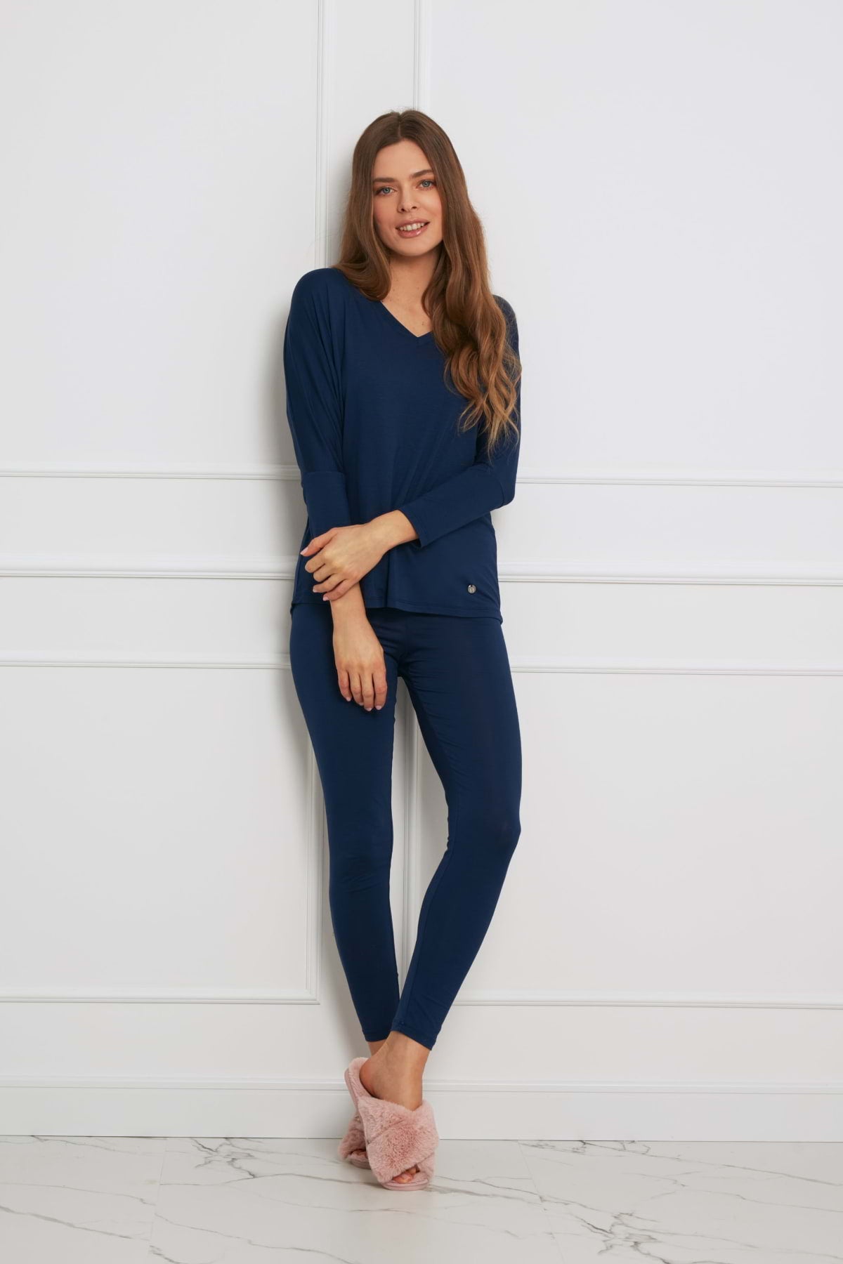 Kobieta z długimi brązowymi włosami uśmiecha się na tle białej ściany w Loungewear wiskozowy komplet z legginsami - granatowy, z bluzką z długim rękawem i pasującymi legginsami, w połączeniu z różowymi rozmytymi kapciami. Jedna noga jest skrzyżowana przed drugą.