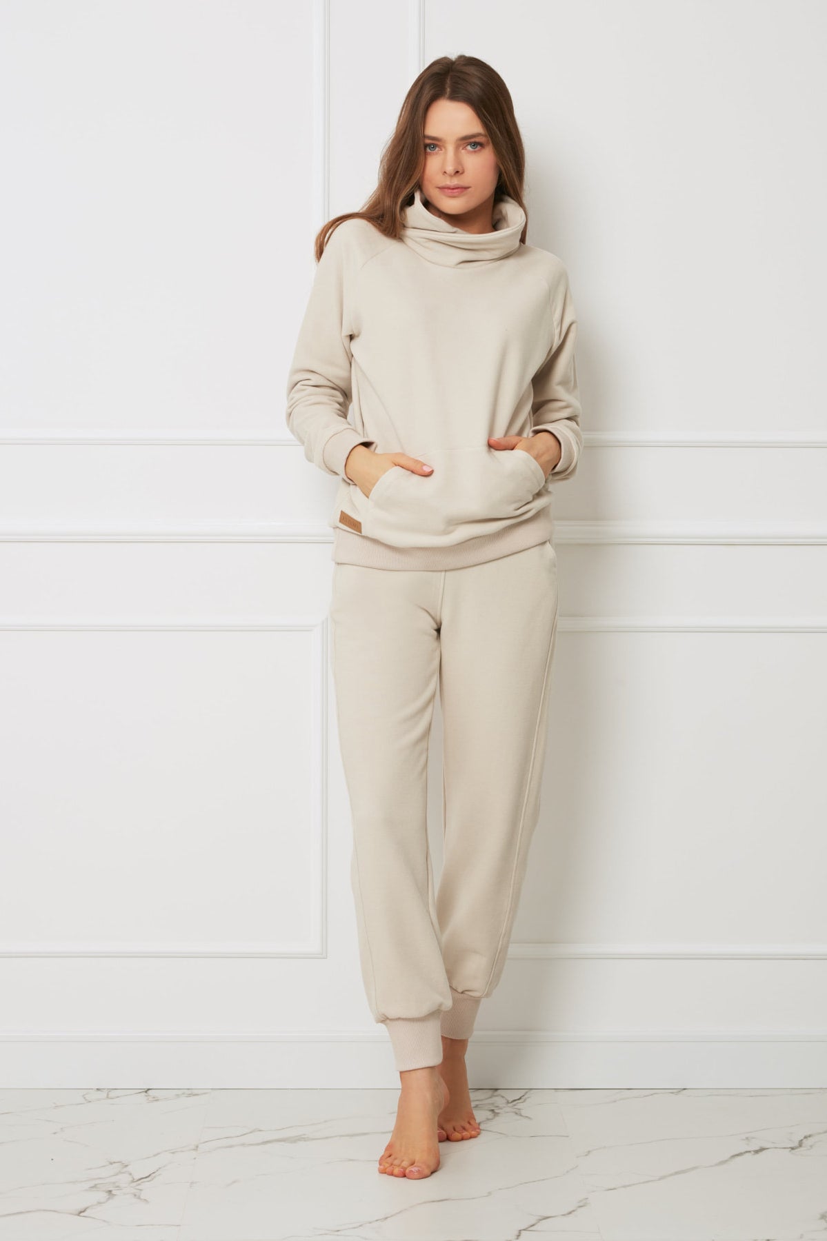 Kobieta stoi boso przy białej ścianie ubrana w jasnobeżową bluzę z kapturem z kieszeniami z przodu i pasujące do niej miękkie spodnie bawełniane Loungewear - beżowe wykonane z dresowych dzianin, wpatrując się w kamerę ze zrelaksowanym wyrazem twarzy.