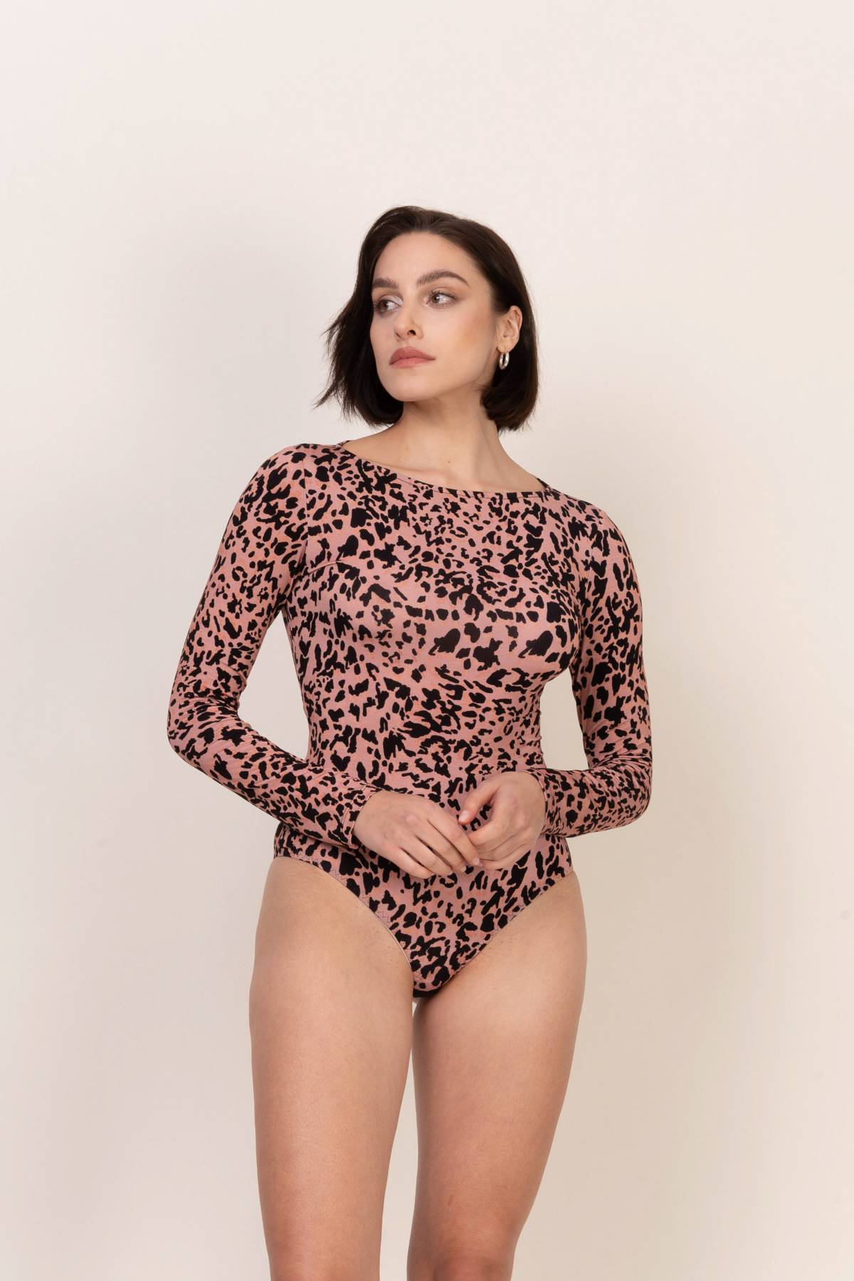 Kobieta o krótkich ciemnych włosach nosi Loungewear wiskozowe body z długim rękawem i dekoltem łódką - panterkę, stojąc na jasnym tle i patrząc lekko w lewo z rękami splecionymi przed sobą.