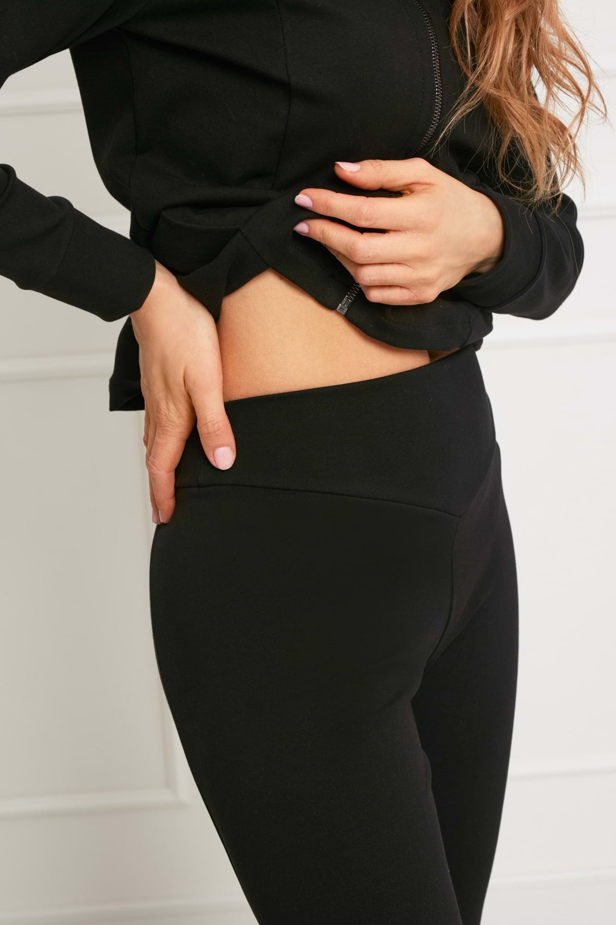 Kobieta stoi na białym tle, podnosząc czarny top z zamkiem błyskawicznym, aby odsłonić wysoki pas Loungewear dopasowane spodnie bawełniane - czarne.