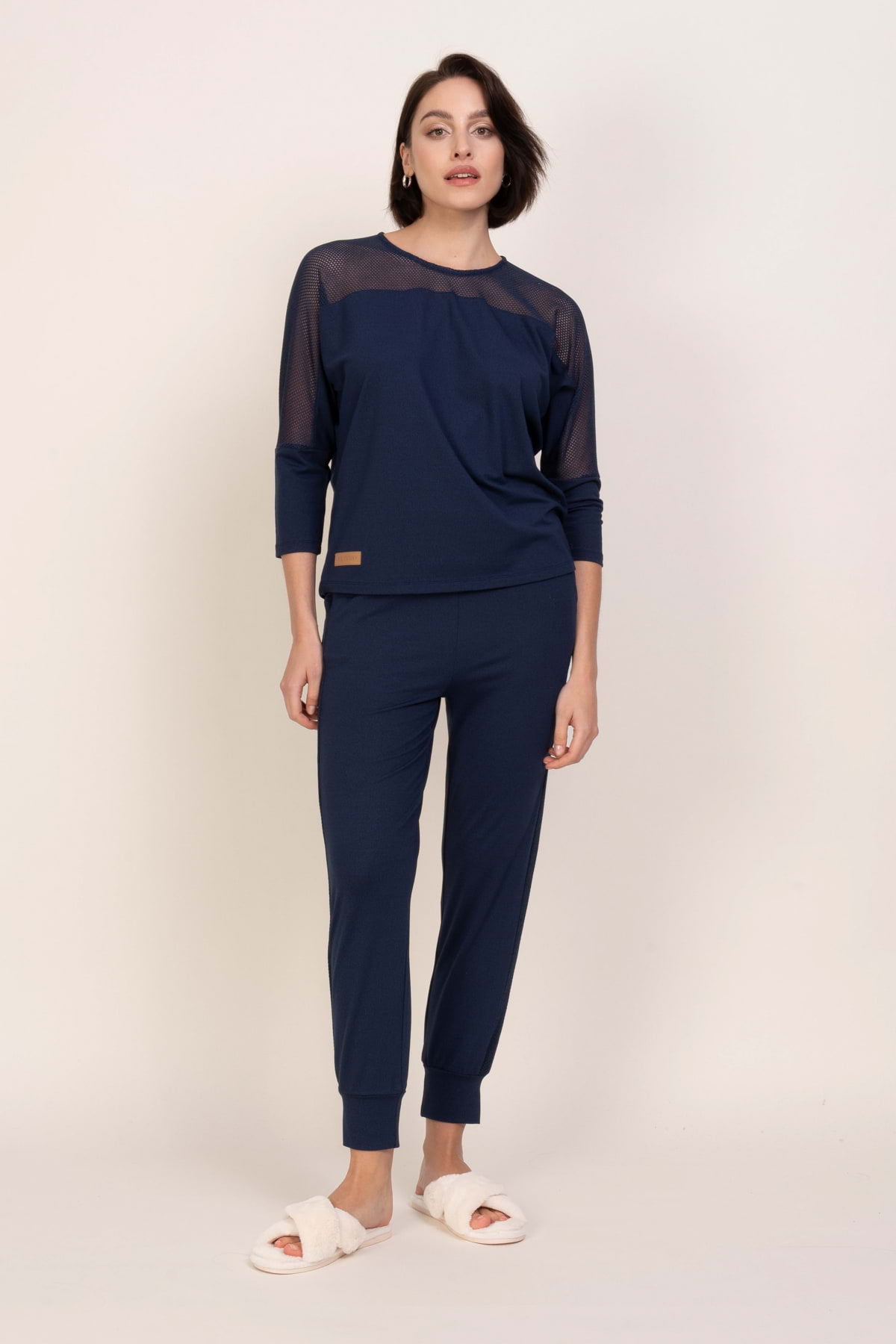 Kobieta z krótkimi brązowymi włosami i neutralnym wyrazem twarzy modeluje zestaw Loungewear wiskozowa bluzka z siateczką - ciemny dżins, składający się z siateczkowego topu i pasujących spodni, na jednolitym jasnym tle. Nosi białe miękkie kapcie.