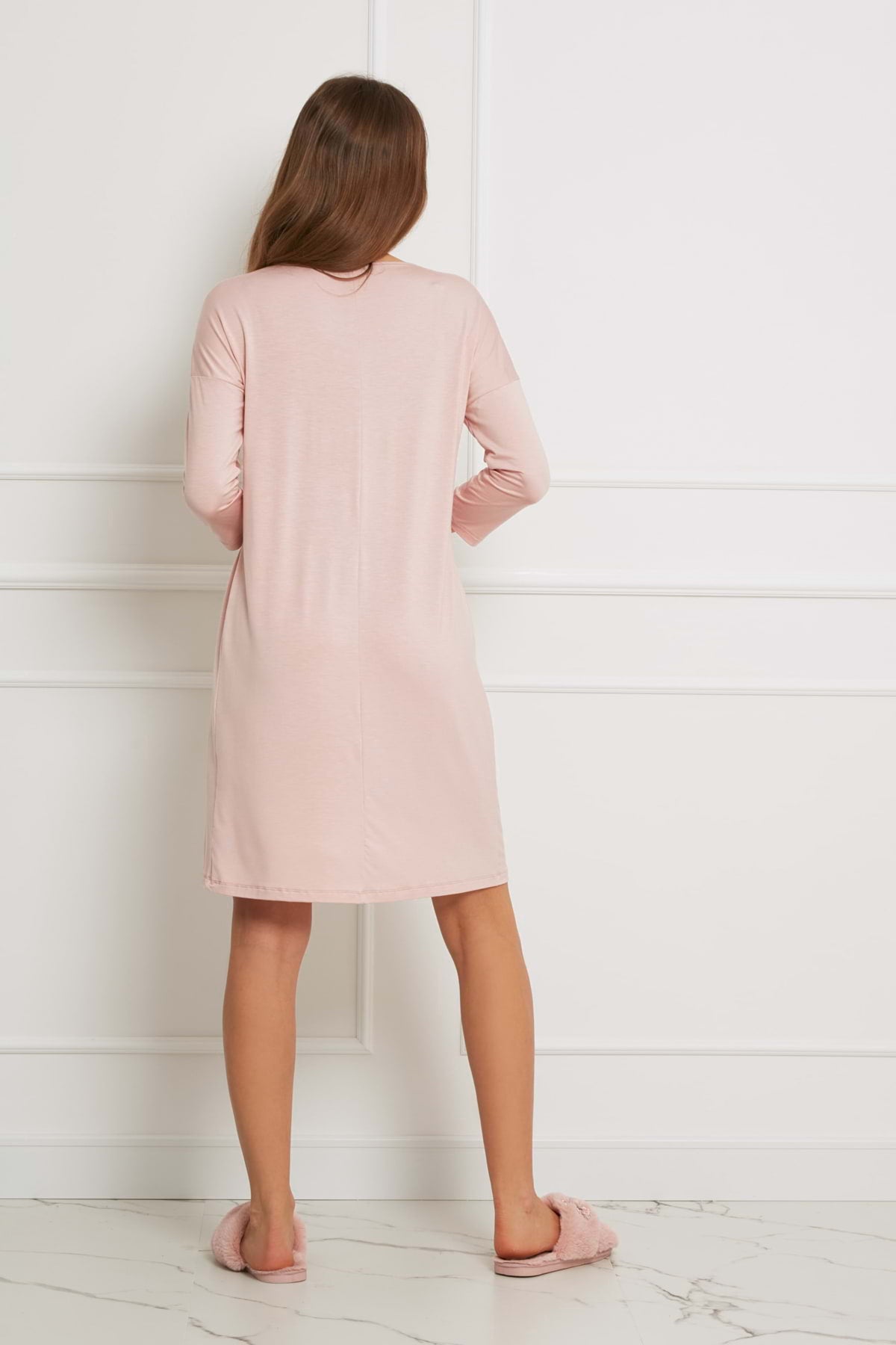 Kobieta stoi w pomieszczeniu, odwrócona tyłem do kamery, ubrana w wiskozową luźną sukienkę Loungewear - brudny róż i różowe kapcie. Pokój ma białe ściany wyłożone panelami i białą marmurową podłogę.