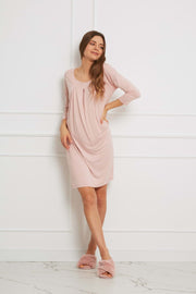 Kobieta w wiskozowej luźnej sukience Loungewear - brudny róż i pasujących do niej różowych kapciach stoi pod białą ścianą, jedną rękę trzymając na biodrze, a drugą u boku, wyglądając na zrelaksowaną.