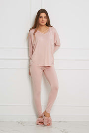 Kobieta z długimi brązowymi włosami stoi na tle białej ściany, ubrana w Loungewear wiskozowy komplet z legginsami - brudny róż, składający się z bluzki z długim rękawem, pasujących legginsów i różowych puszystych kapci. Wygląda na zrelaksowaną z rękami w kieszeniach.