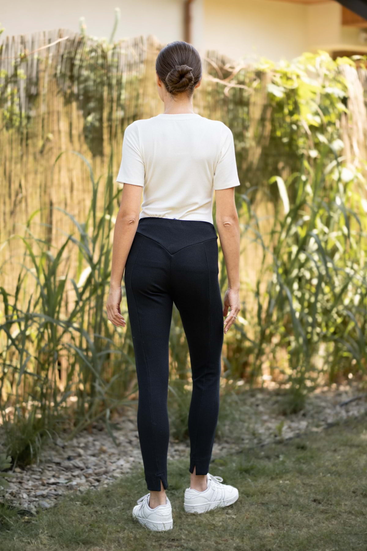 Kobieta z brązowymi włosami spiętymi w kok stoi na zewnątrz odwrócona tyłem, ubrana w Loungewear wiskozowy T-SHIRT w serek - kremowy, czarne legginsy i białe trampki, z zielenią i plecionym płotem w tle.