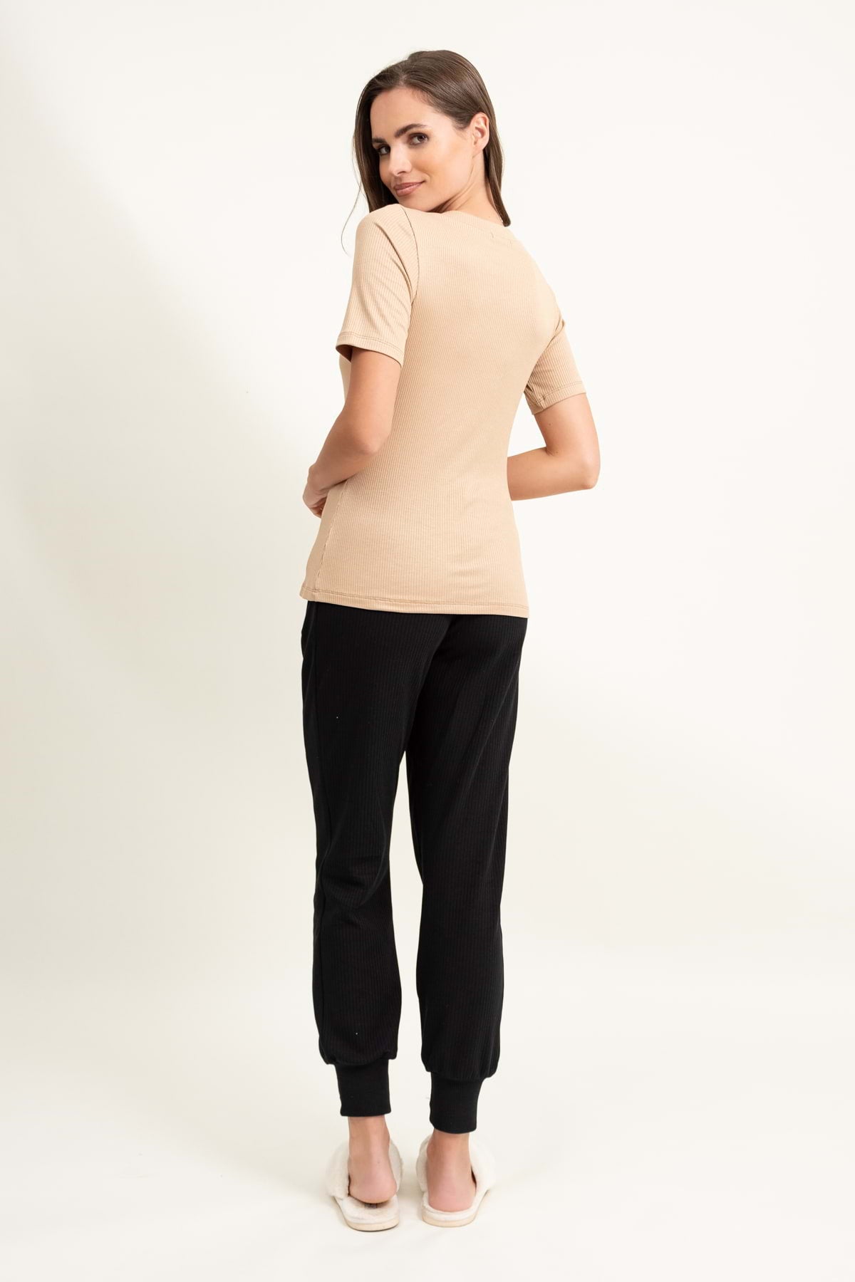 Kobieta odwrócona tyłem, ubrana w Loungewear wiskozowy T-SHIRT w serek - camel, czarne spodnie jogger i białe wsuwane buty. Spogląda przez ramię i uśmiecha się na białym tle.