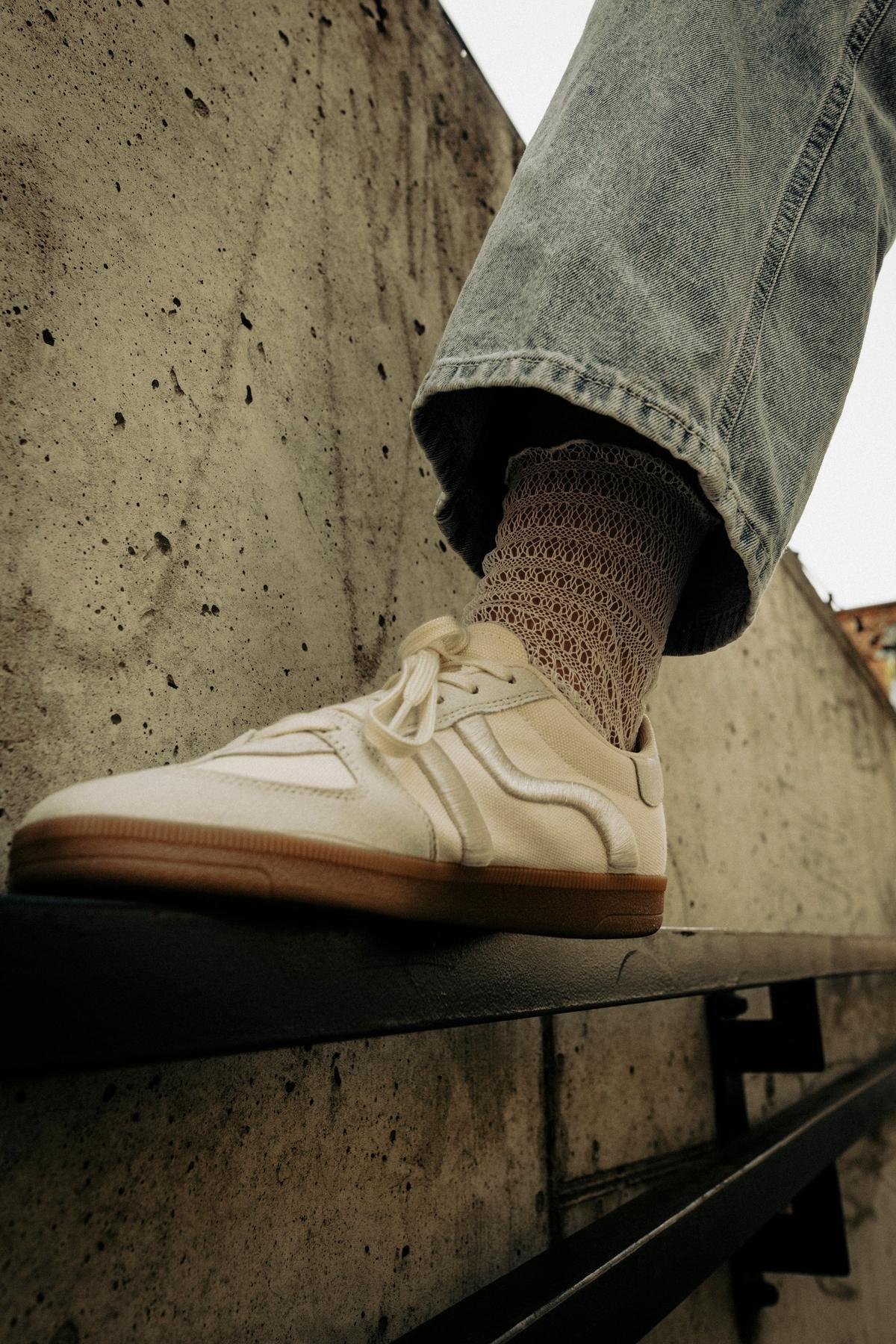 Zbliżenie na stopę w białych trampkach Skarpetki Kabaretki - Waniliowa Sieć i jasnoniebieskich jeansach, stojącą na metalowej barierce przy betonowej ścianie - idealna inspiracja do eleganckich stylizacji.