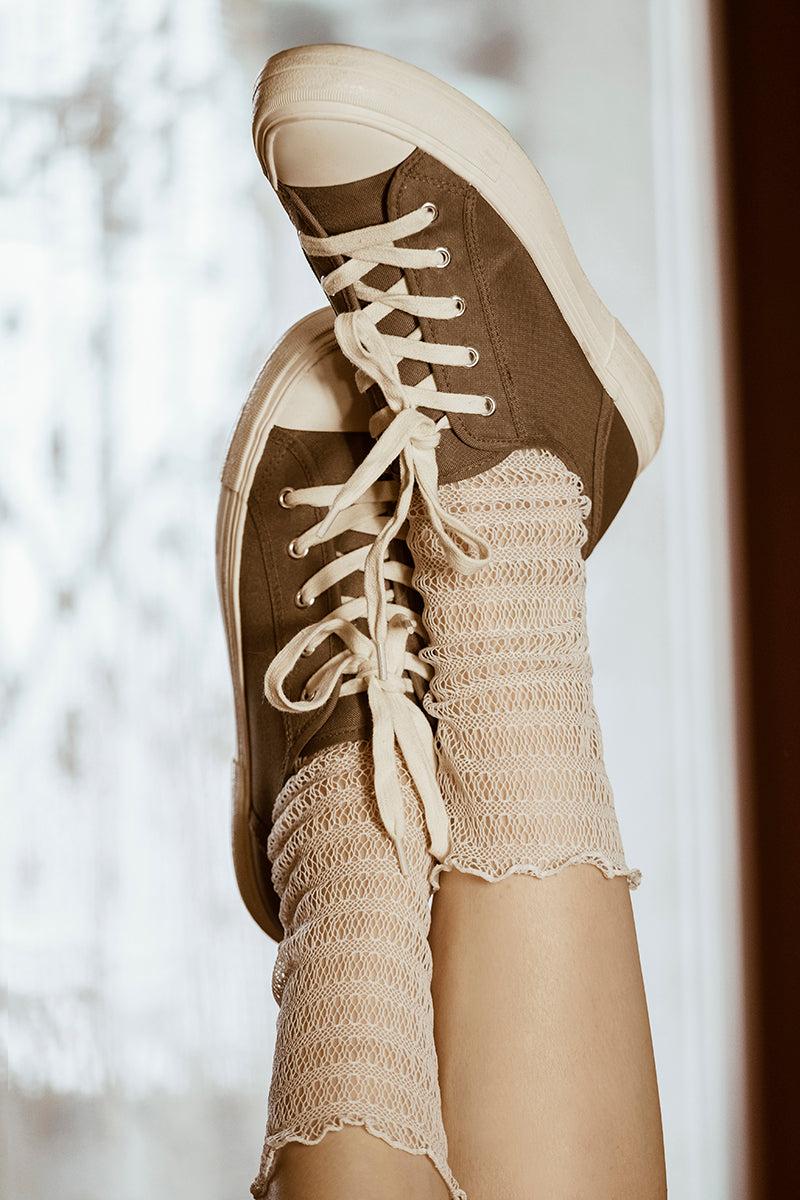 Zbliżenie na dwie uniesione nogi, ubrane w kabaretki Skarpetki - Waniliowa Sieć i brązowe płócienne trampki z białymi sznurówkami i grubą podeszwą. Delikatnie rozmyte tło sprawia, że idealnie nadaje się do eleganckich stylizacji.