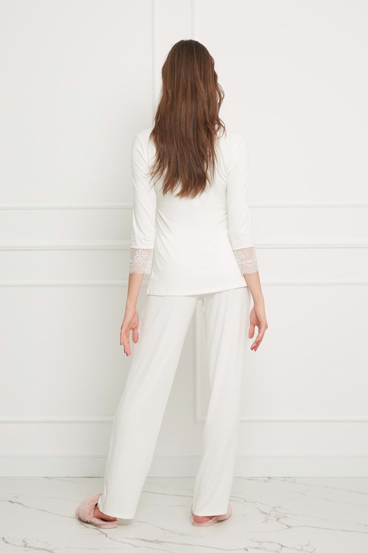 Kobieta z długimi brązowymi włosami stoi boso na marmurowej podłodze, zwrócona plecami do kamery, ubrana w długą piżamę Stars z koronką - kremową i pasujące białe spodnie z koronkowymi mankietami.