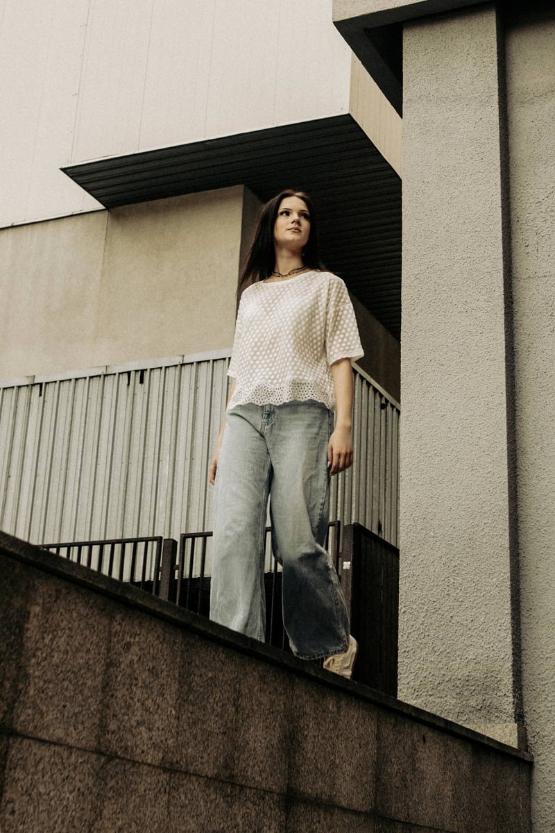 Kobieta ubrana w koszulkę Toscana krótki T-shirt w groszki z koronką - kremowy i jasnoniebieskie dżinsy stoi na miejskiej betonowej konstrukcji, otoczona nowoczesną geometryczną architekturą i patrzy przed siebie z neutralnym wyrazem twarzy.