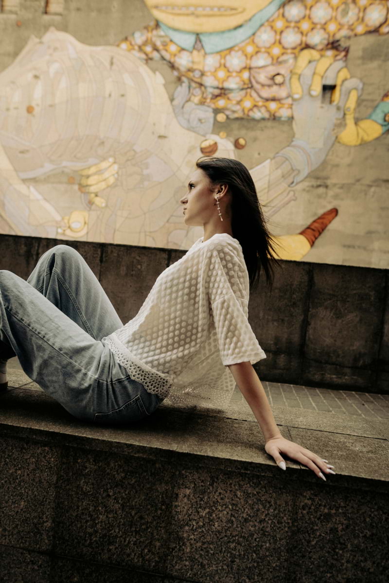 Kobieta w krótkim T-shircie Toscana w groszki z koronką - kremowym i jasnych dżinsach leży na betonowej półce, z kolorowym muralem przedstawiającym dużą postać w tle.