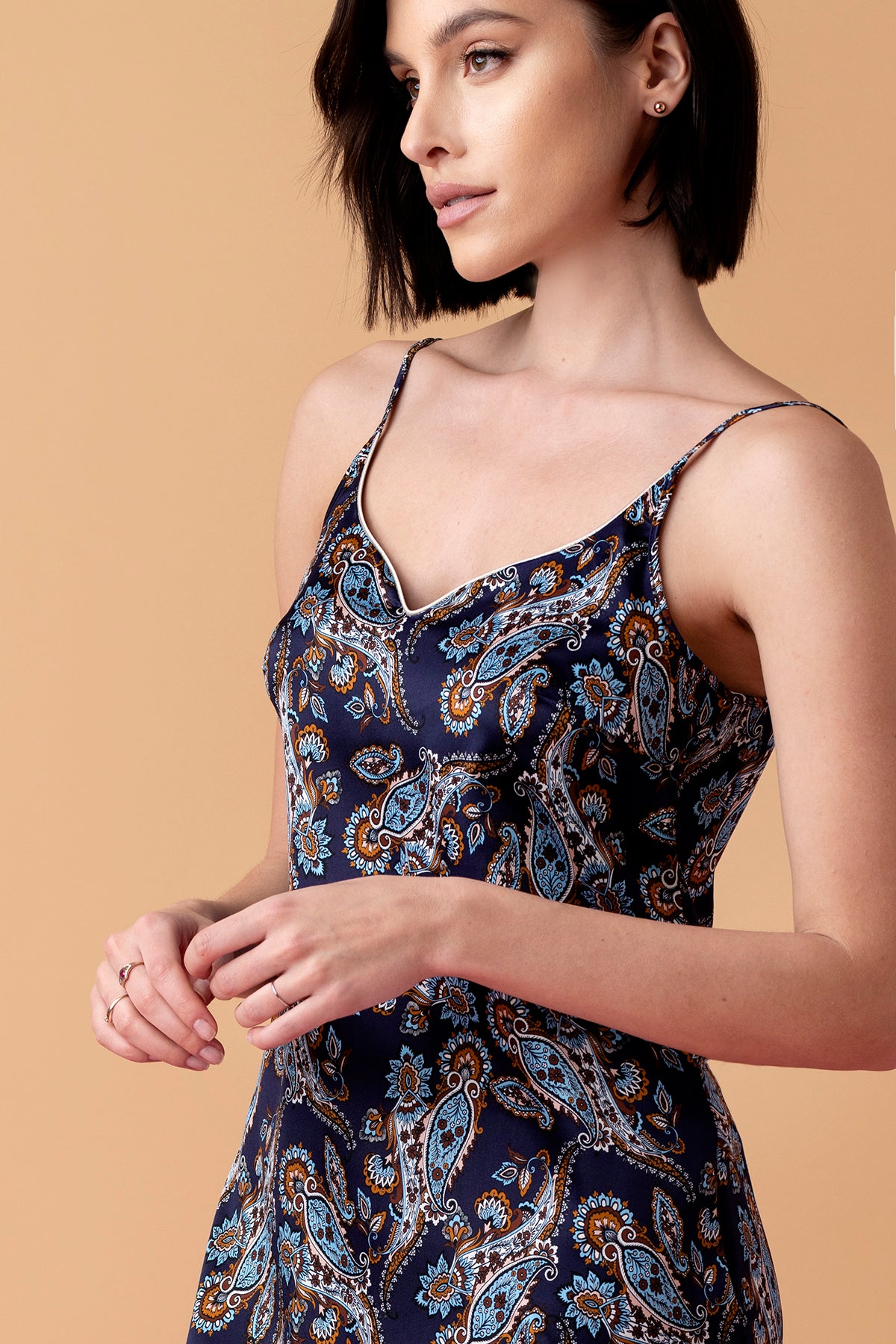 Modelka w granatowej, jedwabnej koszulce na ramiączkach Silk Lullaby z wzorem paisley, długość koszulki przed kolano. Modelka stoi na beżowym tle.