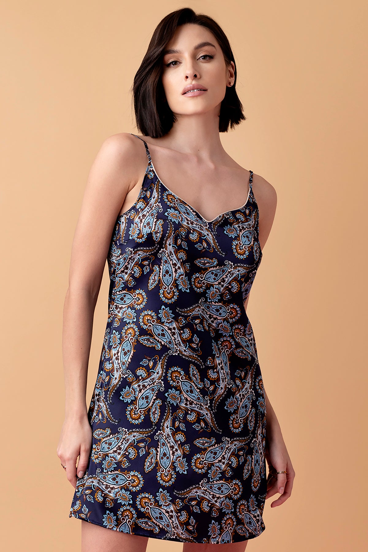 Modelka w granatowej, jedwabnej koszulce na ramiączkach Silk Lullaby z wzorem paisley, długość koszulki przed kolano. Modelka stoi na beżowym tle.