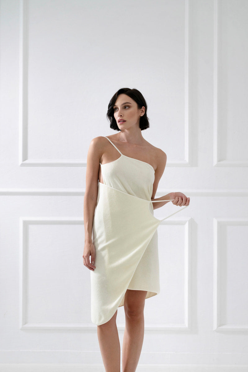 Kobieta o krótkich ciemnych włosach stoi na tle białej ściany wyłożonej panelami, trzymając białą asymetryczną sukienkę. Calma uniwersalna chusta bawełniana na ramiączkach - waniliowa elegancko przewieszona przez ramię.