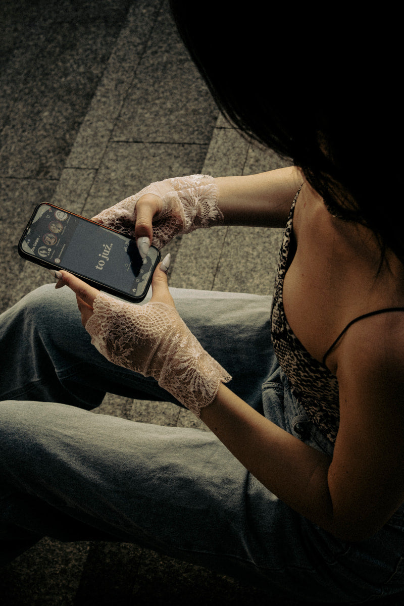 Ubrana w Koronkowe rękawiczki - Pudrowy Dotyk i jeansy osoba siedzi na schodach, trzymając smartfon z otwartą aplikacją muzyczną. Ujęcie z góry podkreśla jej ręce i część tułowia, łącząc romantyzm ze swobodnym stylem.