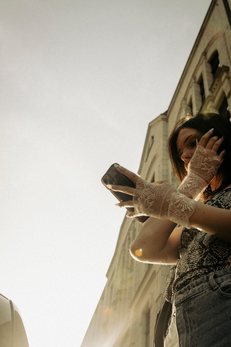 Młoda kobieta w swobodnym stroju i Koronkowe rękawiczki - Pudrowy Dotyk stoi przy wysokim budynku, wpatrując się w telefon z jedną ręką na twarzy. Jasne światło słoneczne dodaje romantycznego stylu temu niskokątnemu ujęciu plenerowemu.