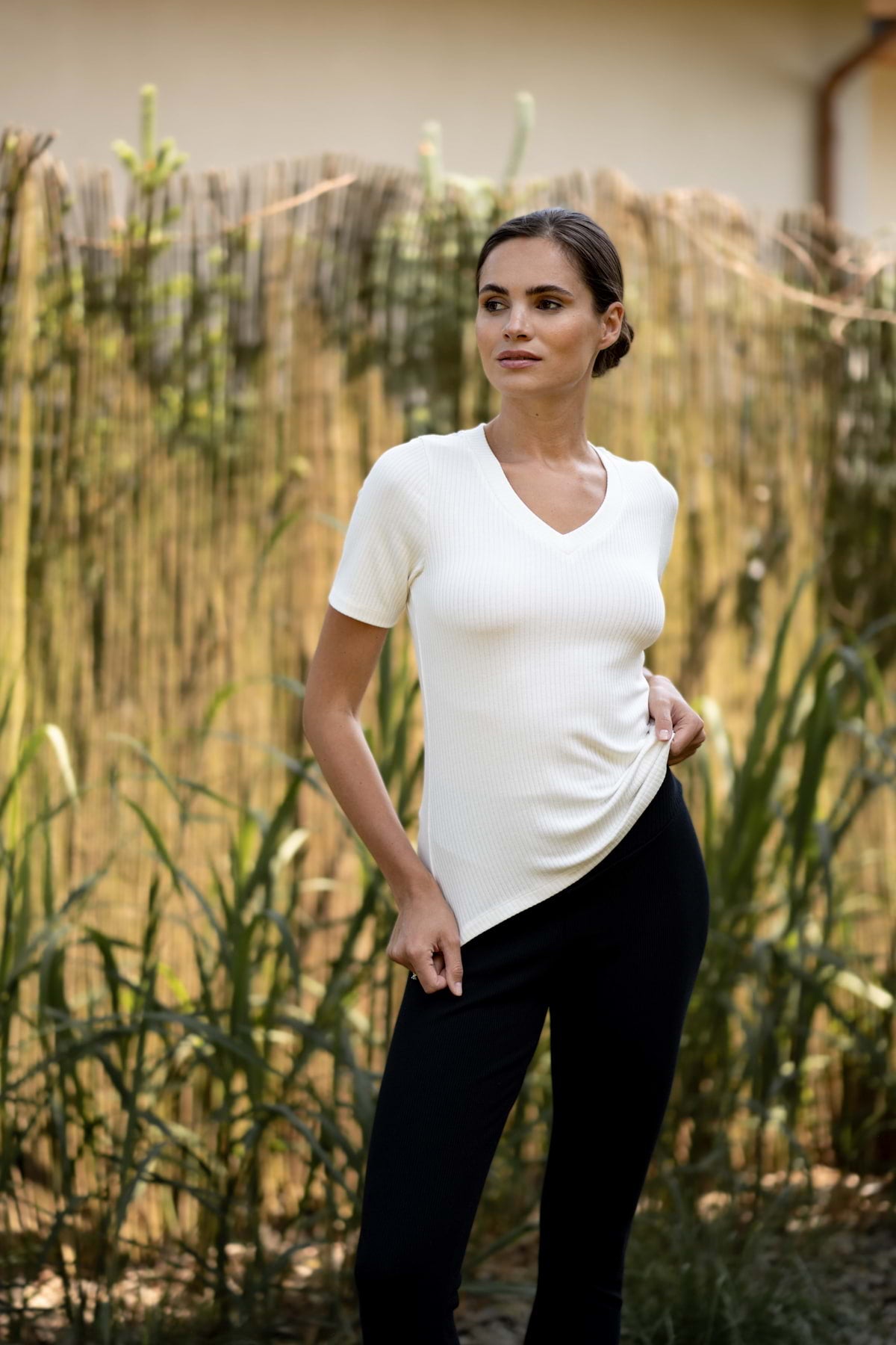 Kobieta stoi na zewnątrz przy wysokiej trawie i bambusowym płocie, ubrana w biały prążkowany top z krótkim rękawem i Loungewear bezuciskowe legginsy prążkowane z karczkiem - czarne, jej włosy są zaczesane do tyłu, a jedna ręka trzyma koszulę.