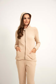 Kobieta ubrana w Loungewear rozpinana bluza z kapturem - jasny beż i pasujące spodnie stoi z rękami w kieszeniach, uśmiechając się lekko na białym tle.