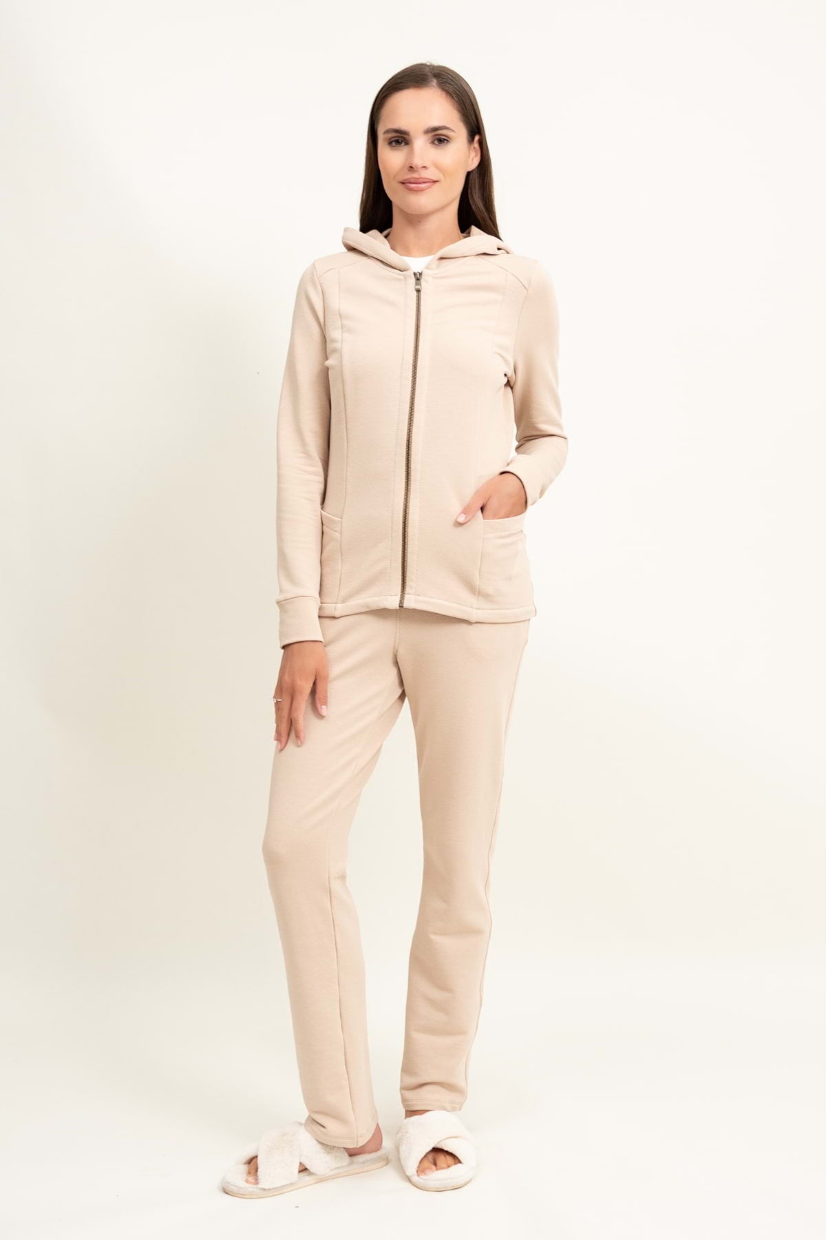 Kobieta stoi na prostym, jasnym tle, ubrana w koszulkę Loungewear rozpinana bluza z kapturem - jasny beż z dopasowanymi spodniami i białymi pantoflami. Uśmiecha się lekko, jedną rękę trzyma w kieszeni, drugą przy boku.