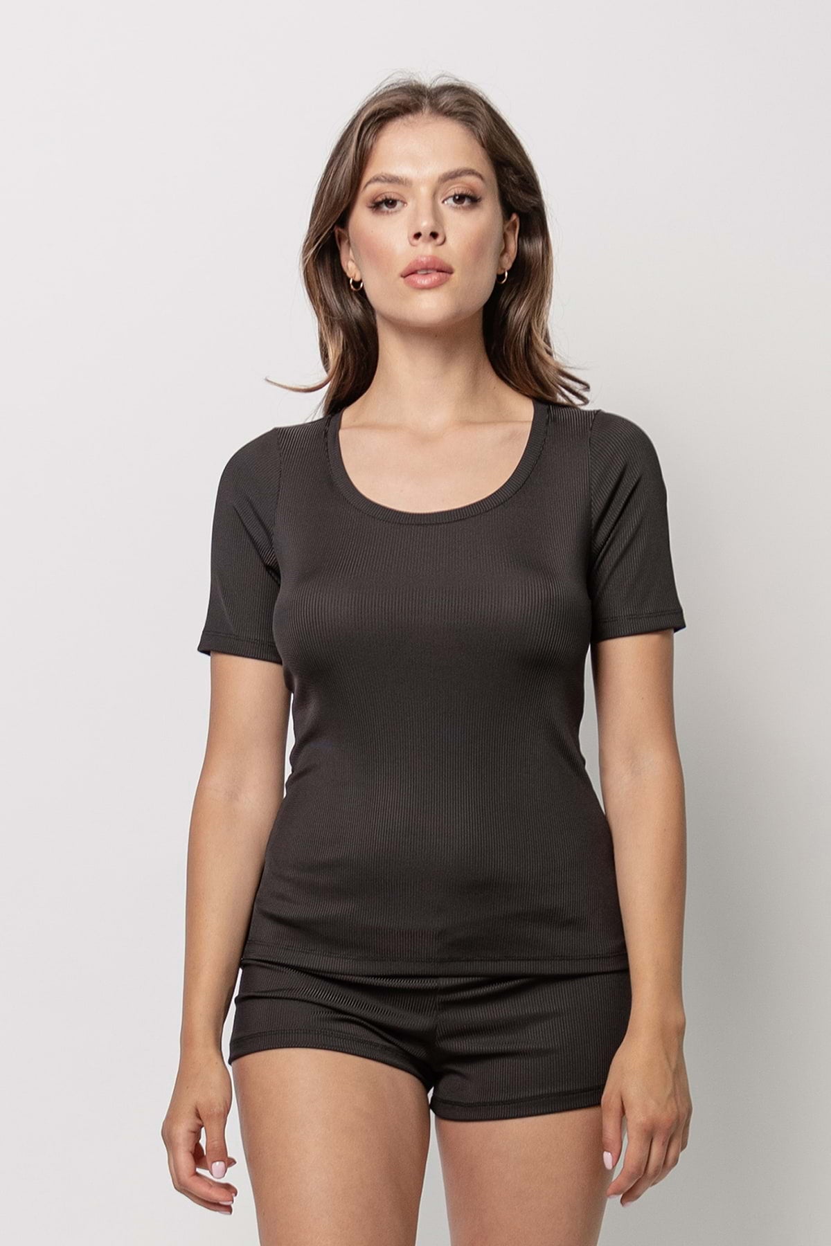 Kobieta stoi na prostym, jasnym tle, ubrana w dopasowaną bluzkę Loungewear z krótkim rękawkiem w prążek - czarną i pasujące czarne szorty. Twarz zwrócona do przodu, ręce rozluźnione po bokach, neutralny wyraz twarzy.