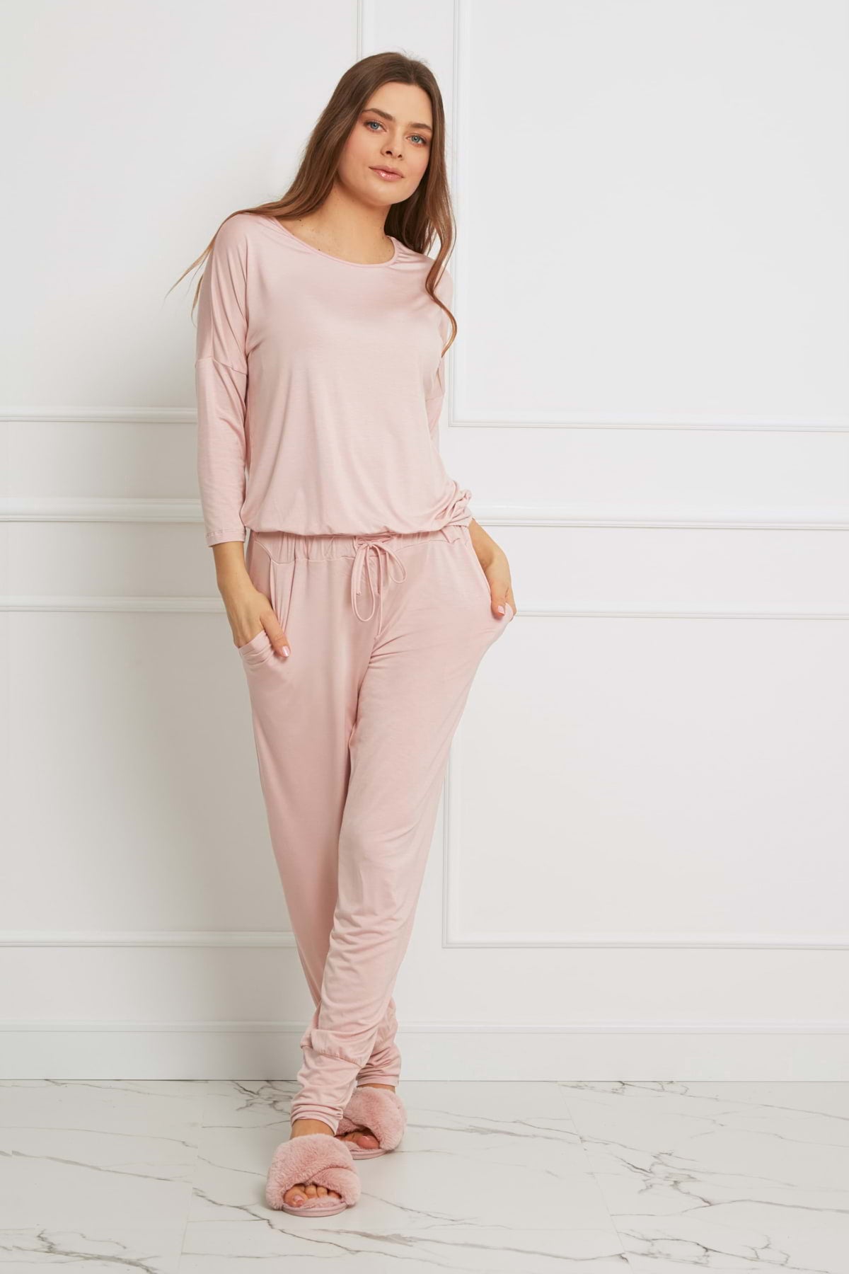 Kobieta o długich brązowych włosach i zrelaksowanym wyrazie twarzy stoi w pomieszczeniu na tle białej ściany, ubrana w Loungewear długi komplet ze spodniami z kieszeniami - brudny róż wykonany z wiskozowej dzianiny, w połączeniu z miękkimi różowymi kapciami.