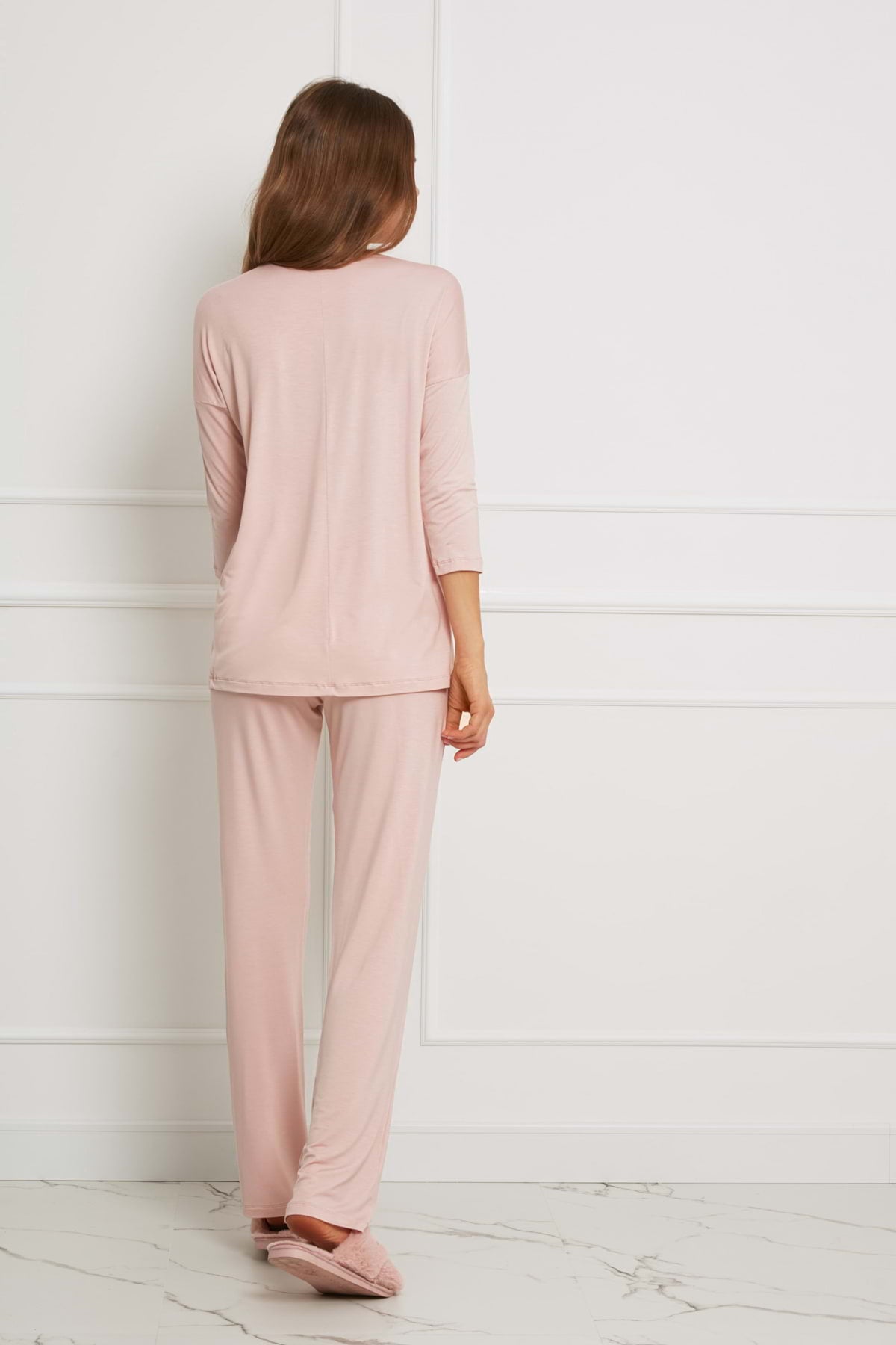 Kobieta z długimi brązowymi włosami stoi naprzeciwko białej ściany, ubrana w Loungewear luźny komplet z długimi spodniami - brudny róż i pasujące kapcie. Jej ręce są rozluźnione po bokach, gdy stoi na białej marmurowej podłodze.
