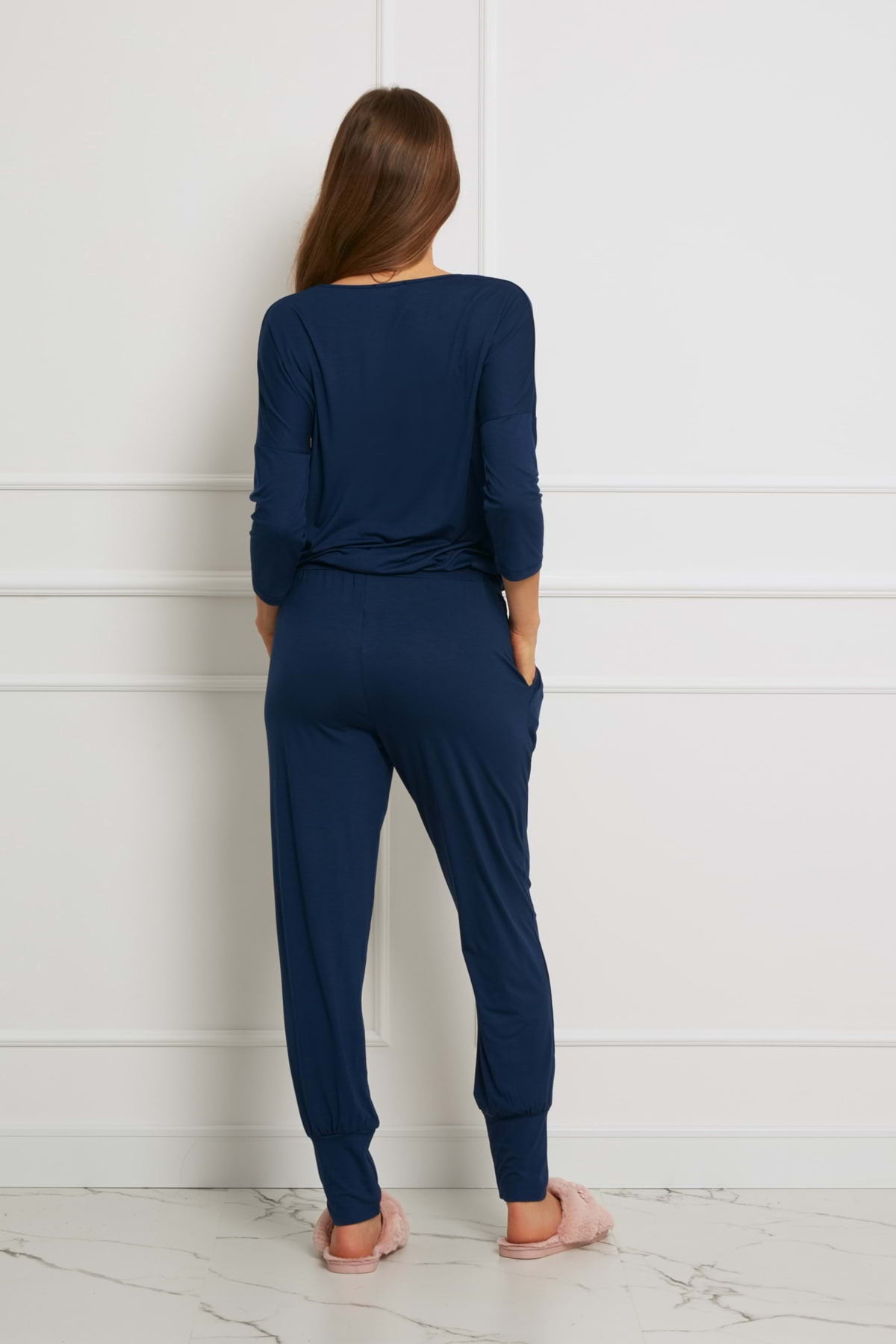 Kobieta stoi przodem do białej ściany, ubrana w Loungewear długi komplet ze spodniami z kieszeniami - granatowy i różowe pantofle. Ukazana jest od tyłu na marmurowej podłodze.